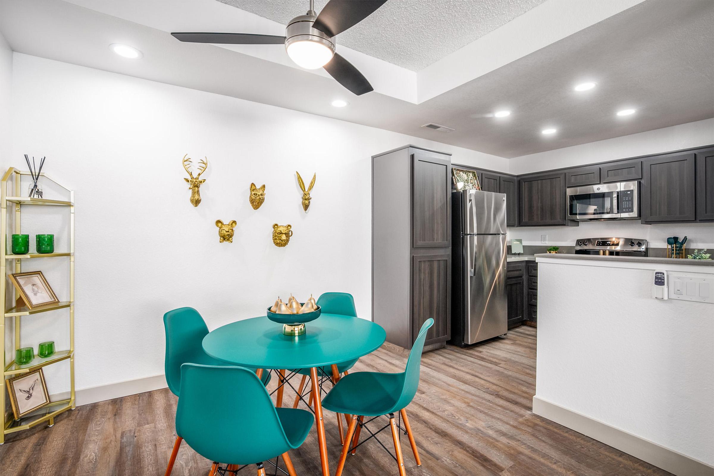 A modern dining area featuring a round teal table with four turquoise chairs. The wall displays decorative animal heads and a gold shelving unit with green glass accents. The kitchen area is visible in the background, showcasing stainless steel appliances and dark cabinetry. A ceiling fan adds a stylish touch.