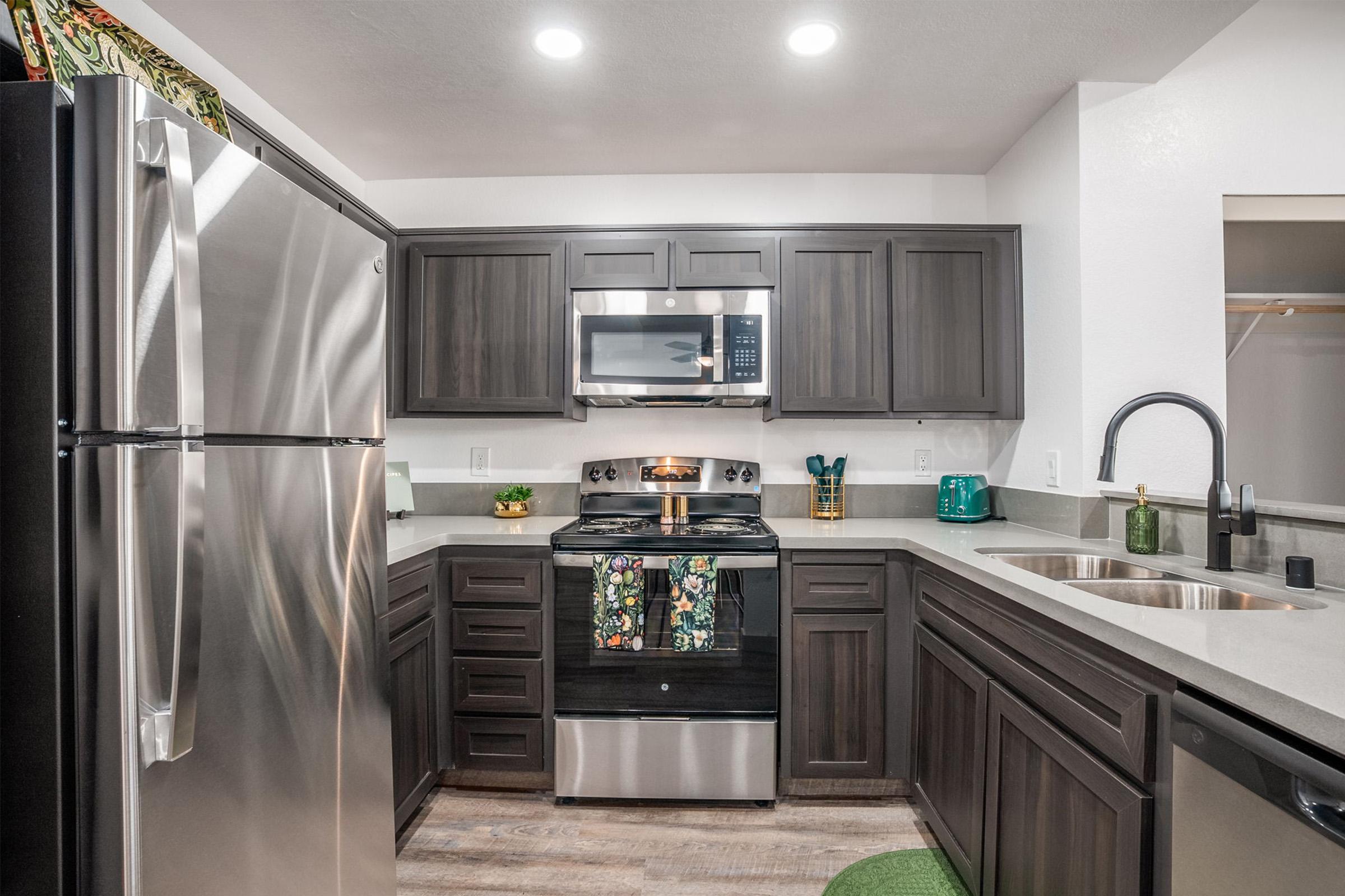 Modern kitchen featuring dark wooden cabinets, stainless steel appliances including a refrigerator, oven, and microwave. A light-colored countertop complements the dark cabinetry, with colorful kitchen towels hanging on the stove. There is a green tea kettle on the counter and a small green stool in the corner.