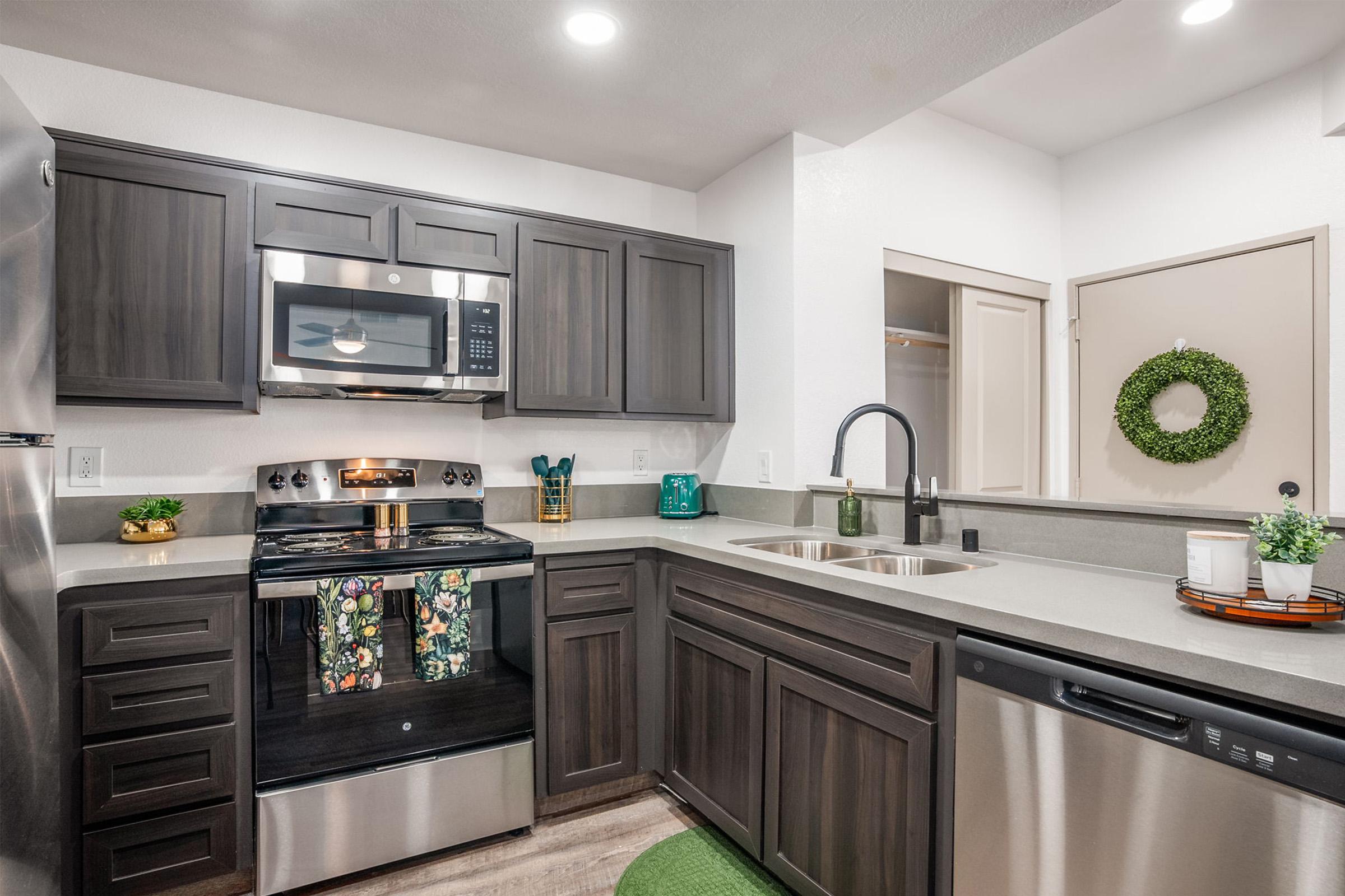 Modern kitchen featuring dark wood cabinets, stainless steel appliances, and a contemporary sink. There is a built-in microwave above the stove, decorative towels on the oven handle, and a stylish green wreath on the wall. The countertop is sleek and light-colored, complementing the overall design.