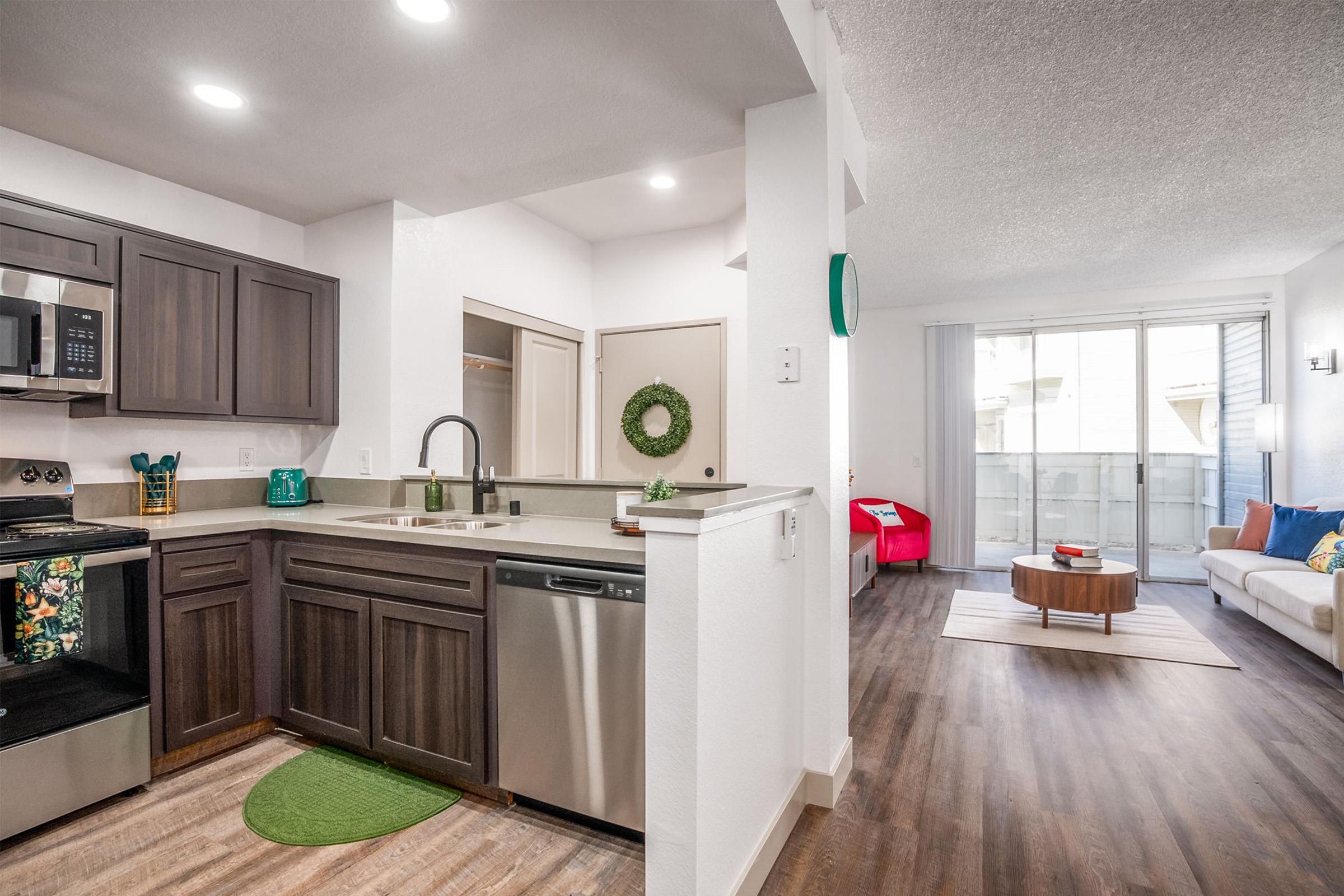 Modern kitchen and living area in an apartment. The kitchen features dark cabinets, stainless steel appliances, and a green area rug. The living area is bright with natural light, showcasing a white couch, decorative pillows, and a round coffee table. A balcony is visible through sliding glass doors.