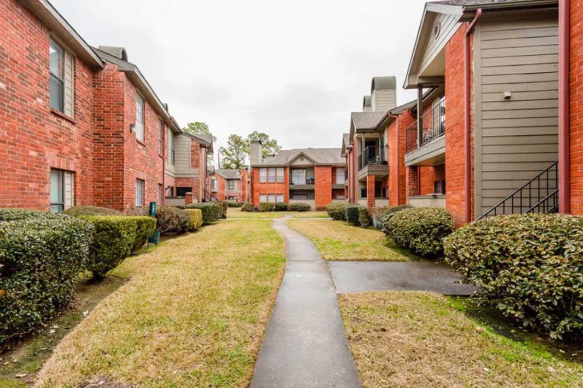 A view of an apartment complex featuring red brick buildings with green landscaping. A paved walkway runs through the grassy area between the buildings, flanked by neatly trimmed hedges. The sky is overcast, creating a subdued atmosphere in the scene.