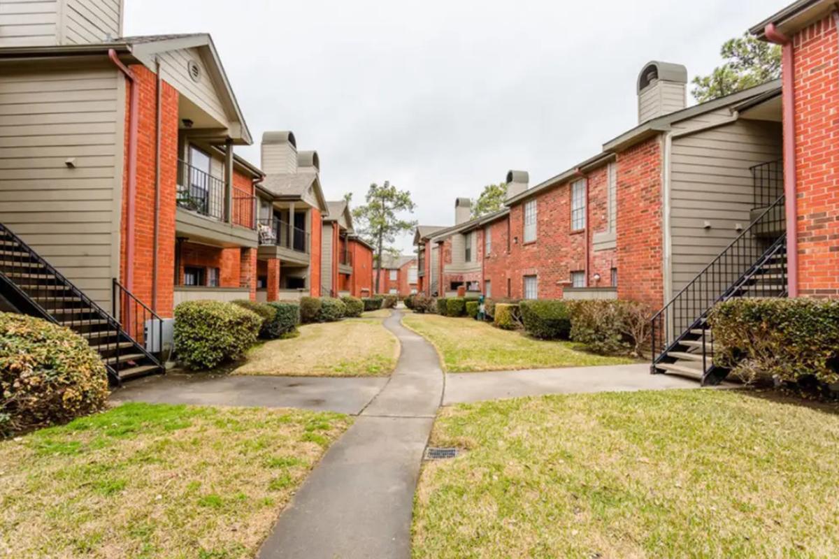 A view of an apartment complex featuring two-story buildings with brick and gray siding, surrounded by manicured lawns and hedges. A paved pathway connects the buildings, and there are staircases leading to the upper units. The sky is overcast, suggesting a cloudy day.