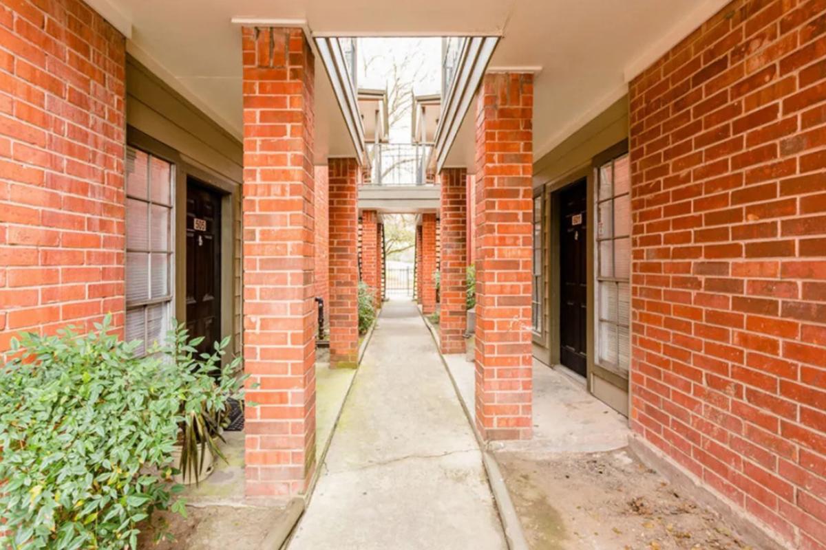 Narrow walkway between brick columns leading to doors on either side. Green shrubs line the path, and natural light filters through an open area above. The scene conveys a sense of quiet and simplicity in an apartment building setting.