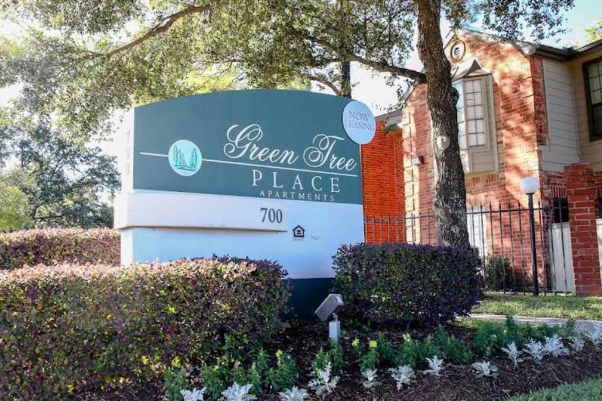 Sign for Green Tree Place Apartments, displaying the name prominently. The sign is set in a landscaped area with low bushes and flower beds. The building in the background has a brick exterior and trees providing shade nearby. The address "700" is visible on the sign.