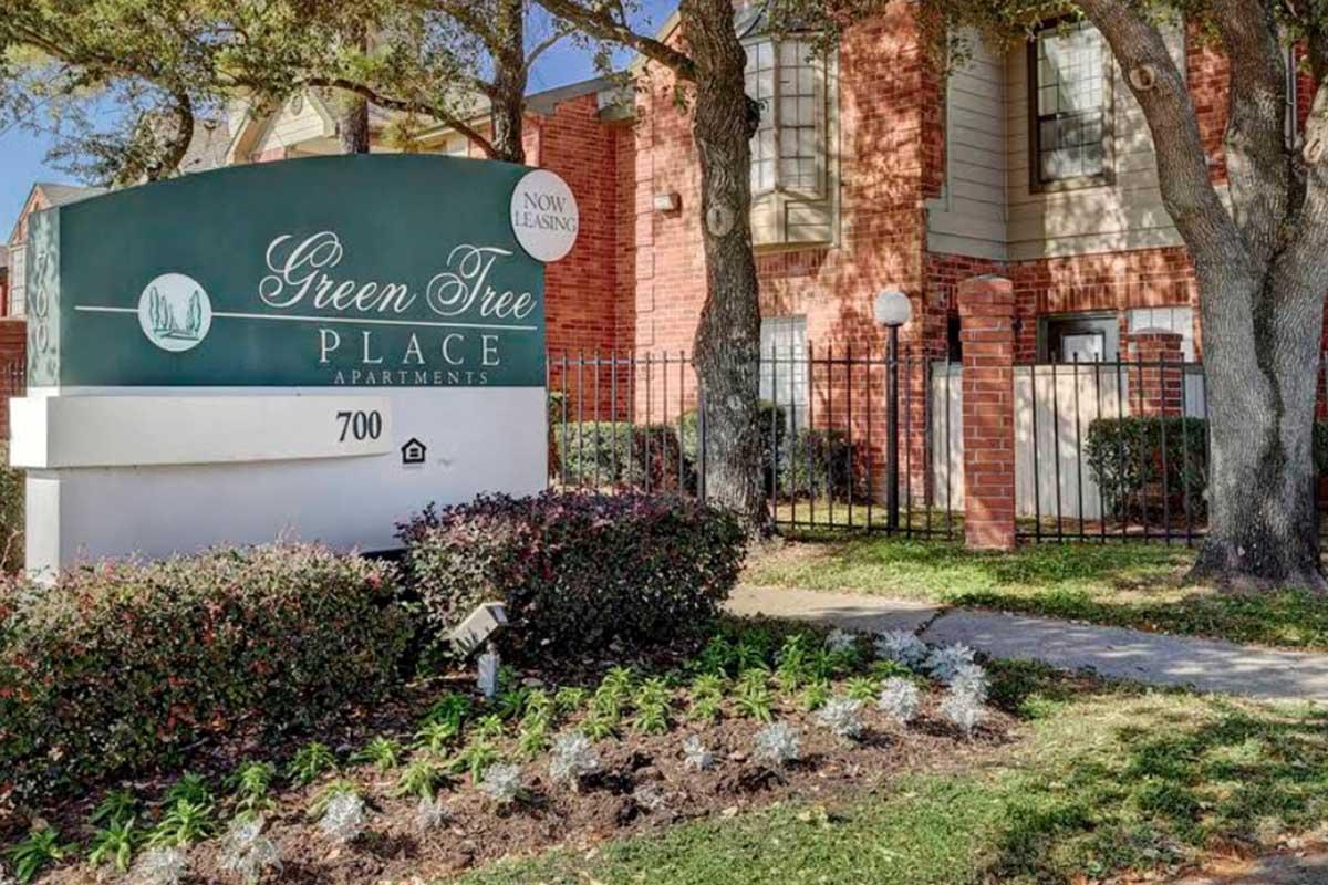 Signage for Green Tree Place Apartments, featuring the name prominently displayed. The sign is surrounded by landscaped areas with shrubs and flowers, with an apartment building visible in the background. The setting includes trees and a gated entrance, conveying a welcoming atmosphere.