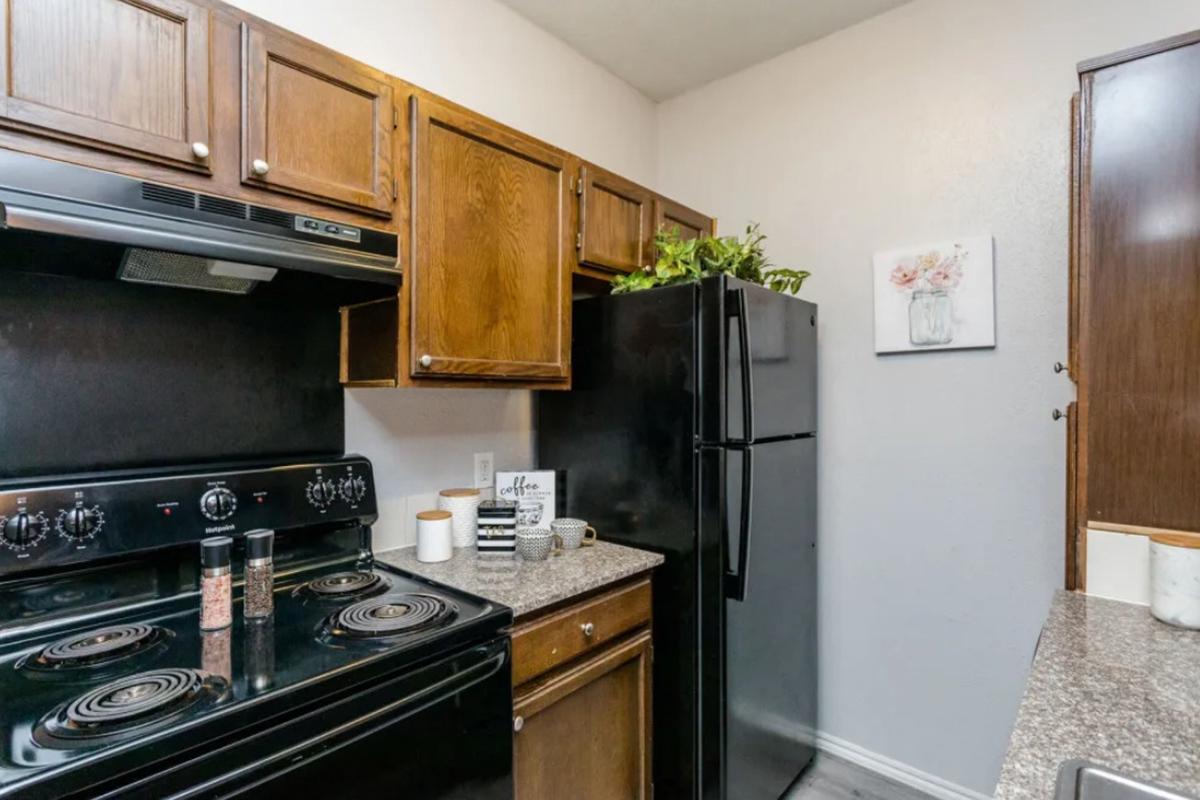 A small kitchen featuring dark wooden cabinetry, a black stove, and a black refrigerator. The countertops are granite, and there are decorative items, including spice jars and a plant. A framed floral picture adorns the wall, adding a touch of color. The space is tidy and well-organized.