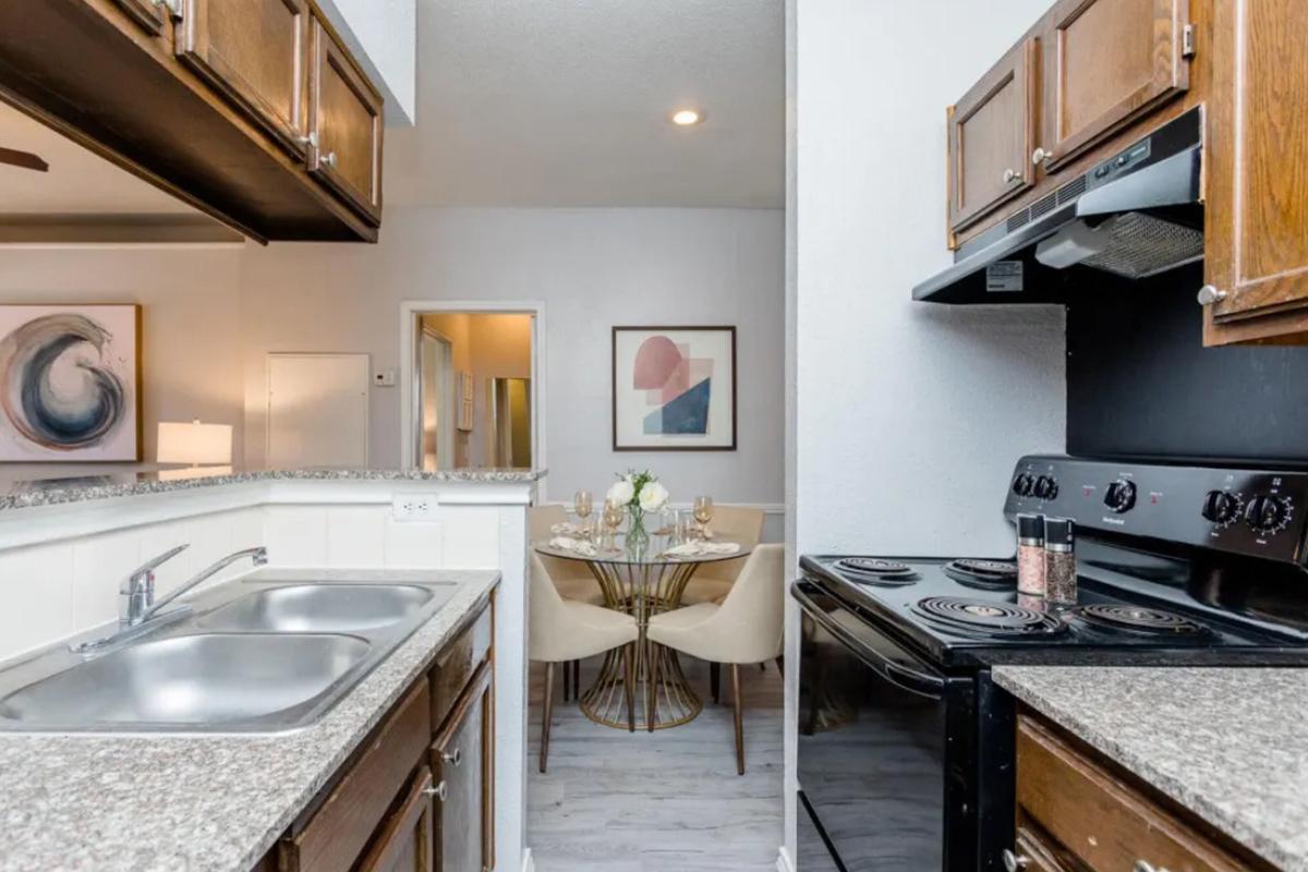 A modern kitchen featuring dark wood cabinets, a black stove, and a double sink with granite countertops. In the background, a dining area with a round table set for four and contemporary art on the walls. The space is well-lit with a neutral color palette, creating a cozy atmosphere.