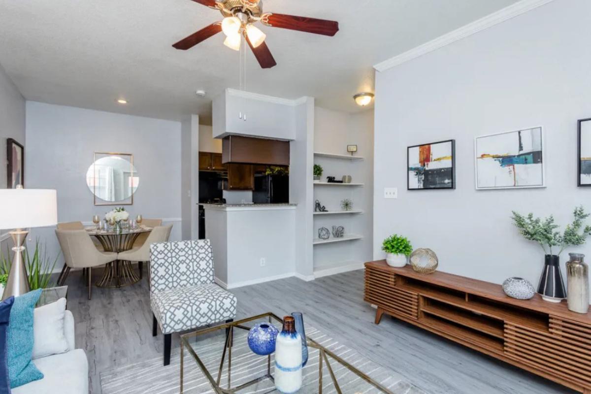 A modern living room featuring a patterned armchair and a wooden coffee table with decorative items. There is a ceiling fan above, and a dining area can be seen in the background. The walls are painted light gray, and artwork adorns the walls, creating an inviting atmosphere with natural light.