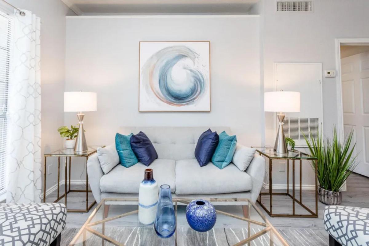 Stylish living room featuring a light gray sofa with blue and teal decorative pillows, two modern lamps on either side, and a decorative coffee table with vases. A piece of abstract wall art above the sofa and potted plants add a fresh touch to the contemporary decor. Bright lighting enhances the open feel of the space.