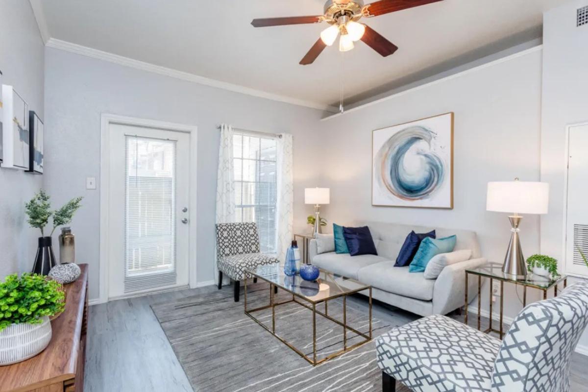 Bright and modern living room featuring a light gray sofa adorned with blue and green accent pillows. Two patterned chairs flank a glass coffee table, while a large piece of wall art adds a stylish touch. A ceiling fan provides airflow, and a door leads to an outdoor area. Natural light floods through large windows.