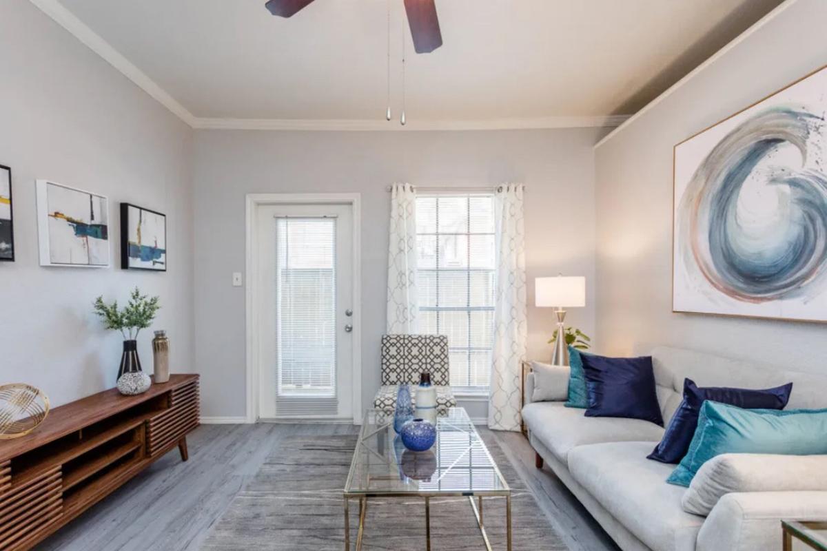 A stylish living room featuring a light gray color scheme, a cream sofa adorned with blue and teal pillows, a glass coffee table, and a wooden media console. There is a wall art piece depicting a swirl design, a potted plant on the console, and large windows with sheer curtains allowing natural light in.