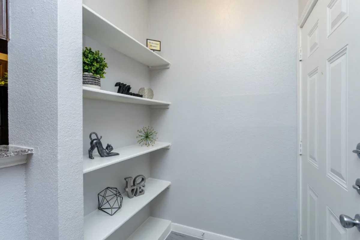 A small nook with white shelves displaying decorative items such as a green plant, black sculptures, and geometric shapes. The walls are light-colored, creating a bright and inviting atmosphere, with a closed door visible in the background.