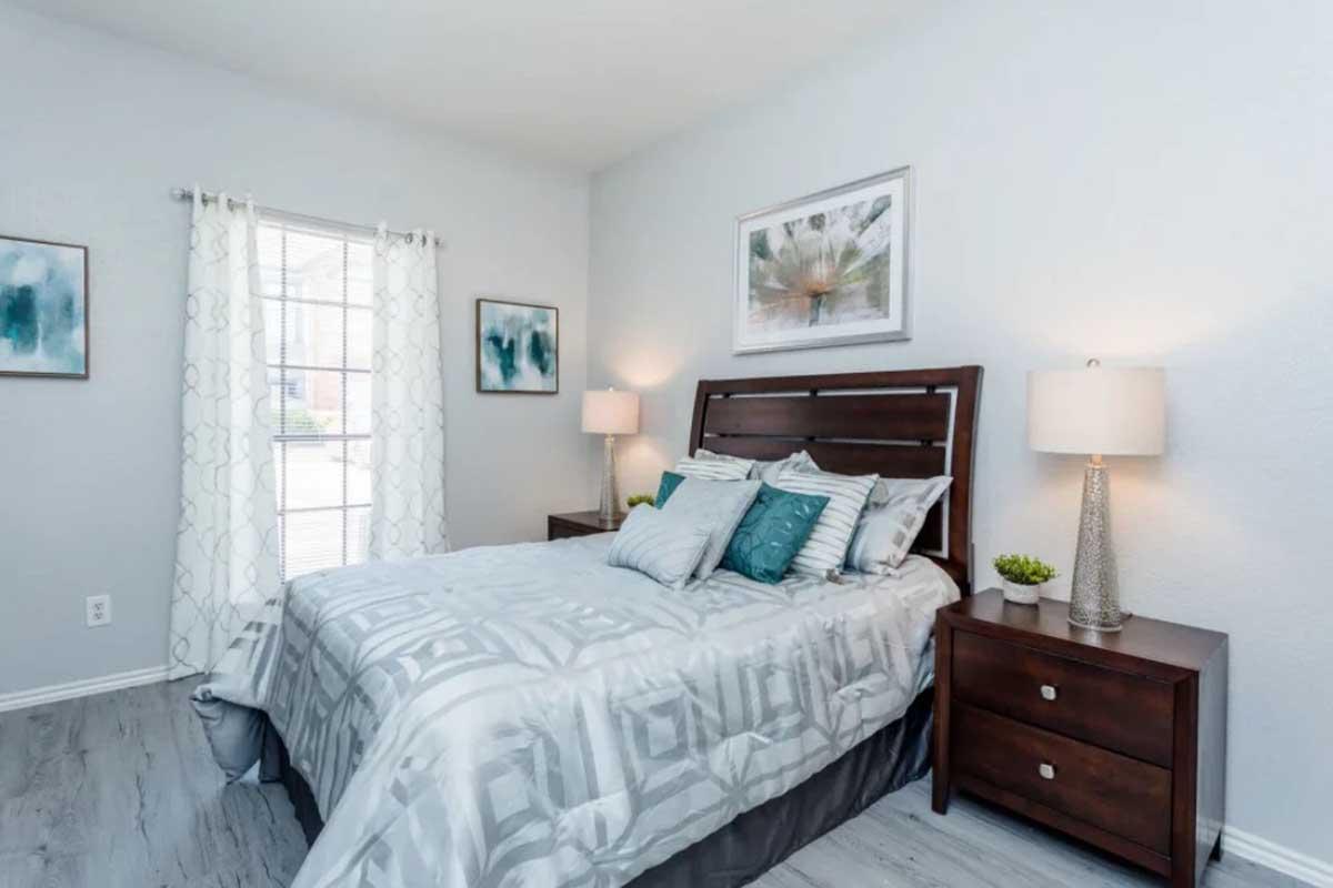 A cozy bedroom featuring a queen-sized bed with gray bedding and decorative pillows. There are two bedside tables with lamps on either side, a window with sheer curtains allowing natural light, and a simple wall art piece above the bed. The room has a light, neutral color palette and wooden flooring.