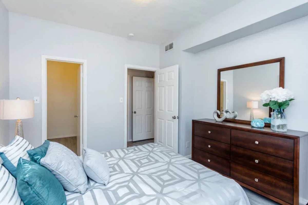 A cozy bedroom featuring light gray walls and a patterned bedspread. The bed is adorned with decorative pillows. A wooden dresser is positioned against the wall, with a mirror above it and decorative items like flowers and ornaments. Two doors lead to adjacent rooms. Soft lighting creates a welcoming atmosphere.