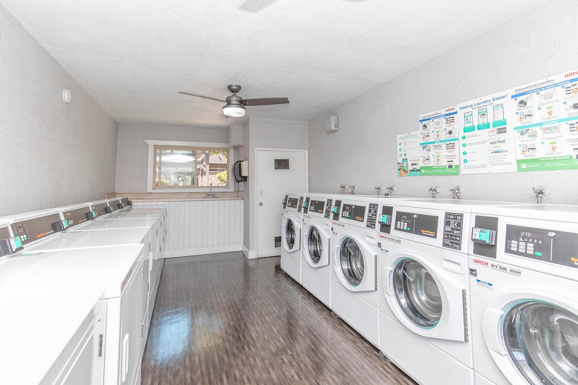 A clean and bright laundry room featuring multiple white washing machines and dryers lined up against the wall. A ceiling fan is visible, and there's a window letting in natural light. An information chart about laundry equipment is mounted on the wall, providing utility instructions.