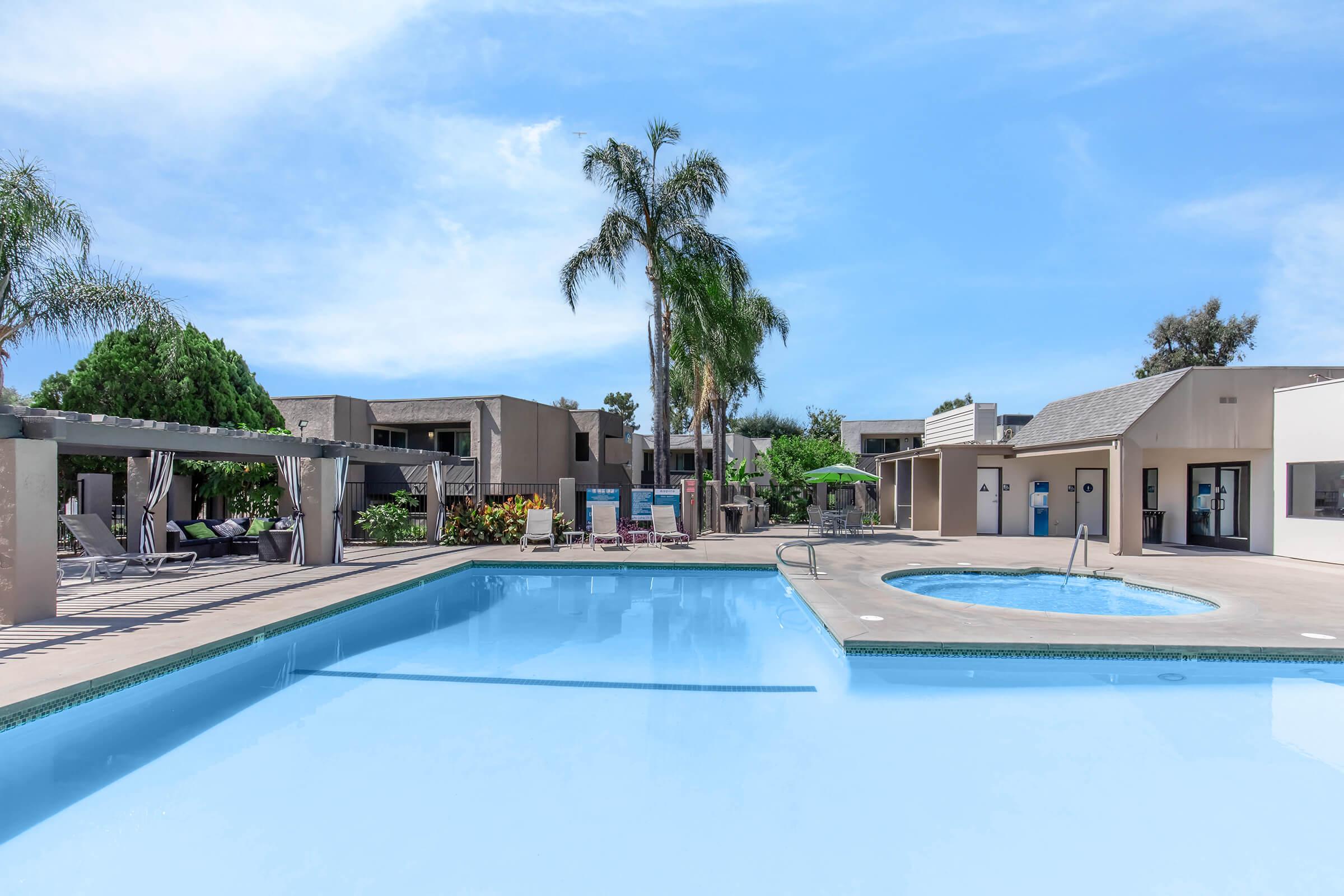 Swimming pool area featuring clear blue water, surrounded by palm trees and lounge chairs. In the background, there are several residential buildings with a sunny sky overhead. A hot tub is visible next to the pool, and there are umbrellas providing shade.
