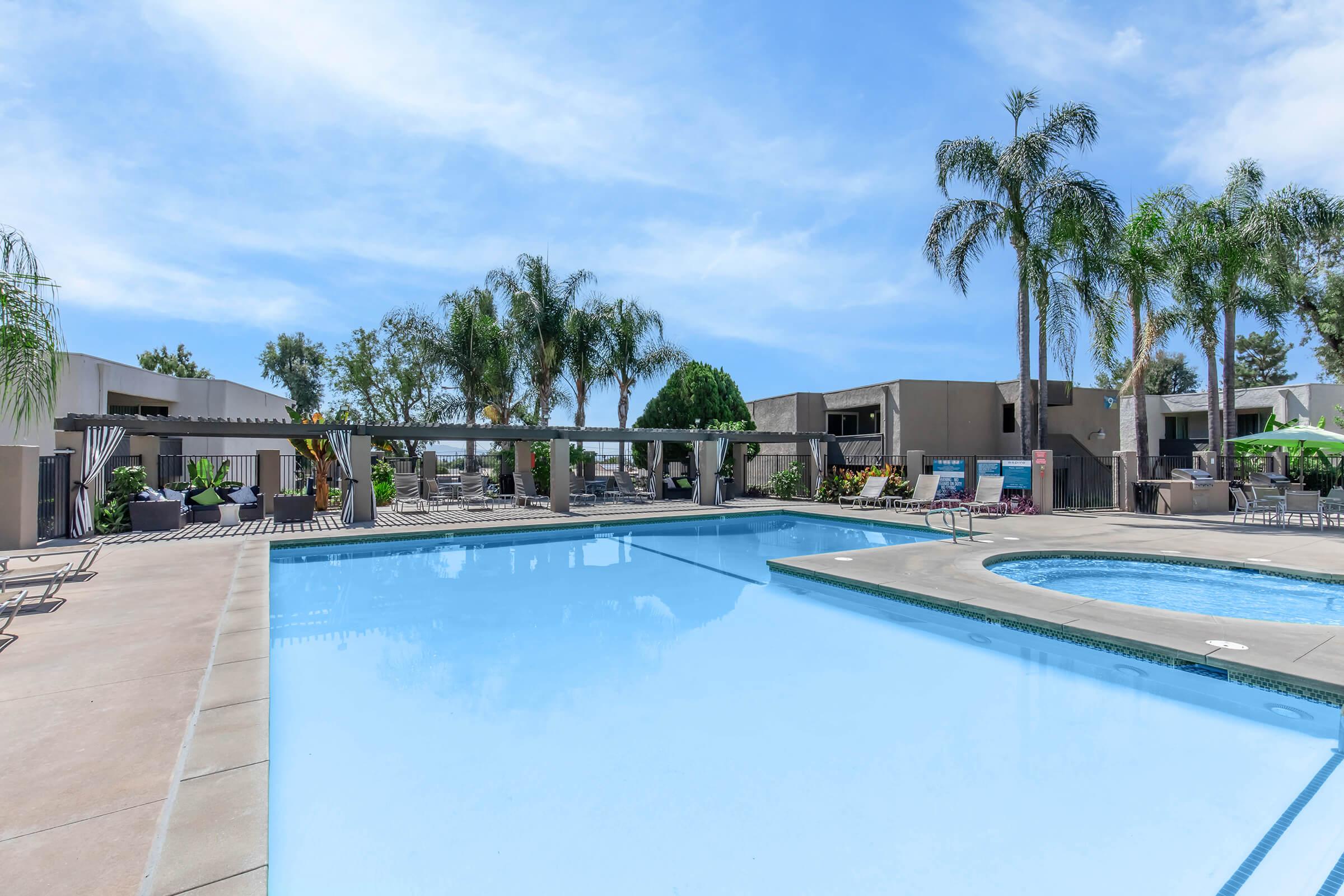 A clear blue swimming pool surrounded by palm trees and lounge chairs, with a smaller hot tub on the side. Modern buildings are visible in the background, and the sky is bright with a few clouds, indicating a sunny day. The setting has a relaxed and inviting atmosphere.