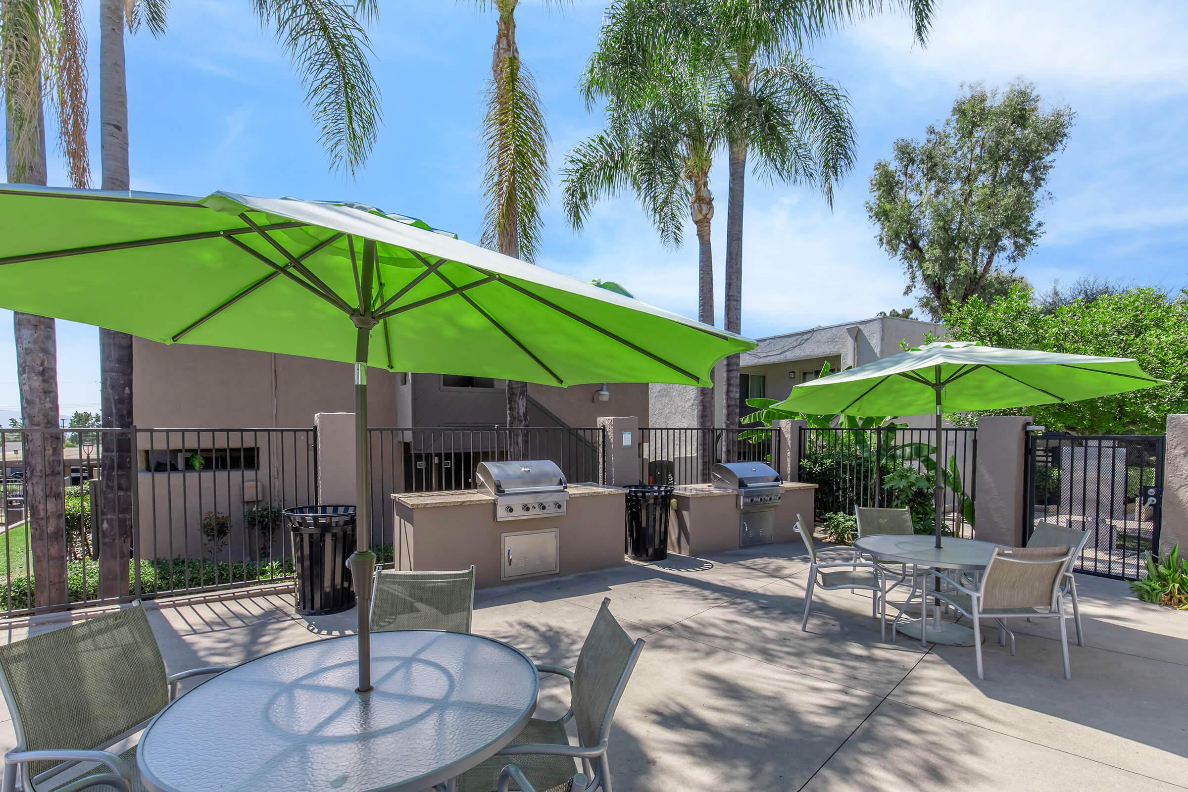 Outdoor patio area with green umbrellas over tables, surrounded by palm trees. Two barbecue grills are visible near the fencing, creating a comfortable setting for dining and socializing in a sunny environment.
