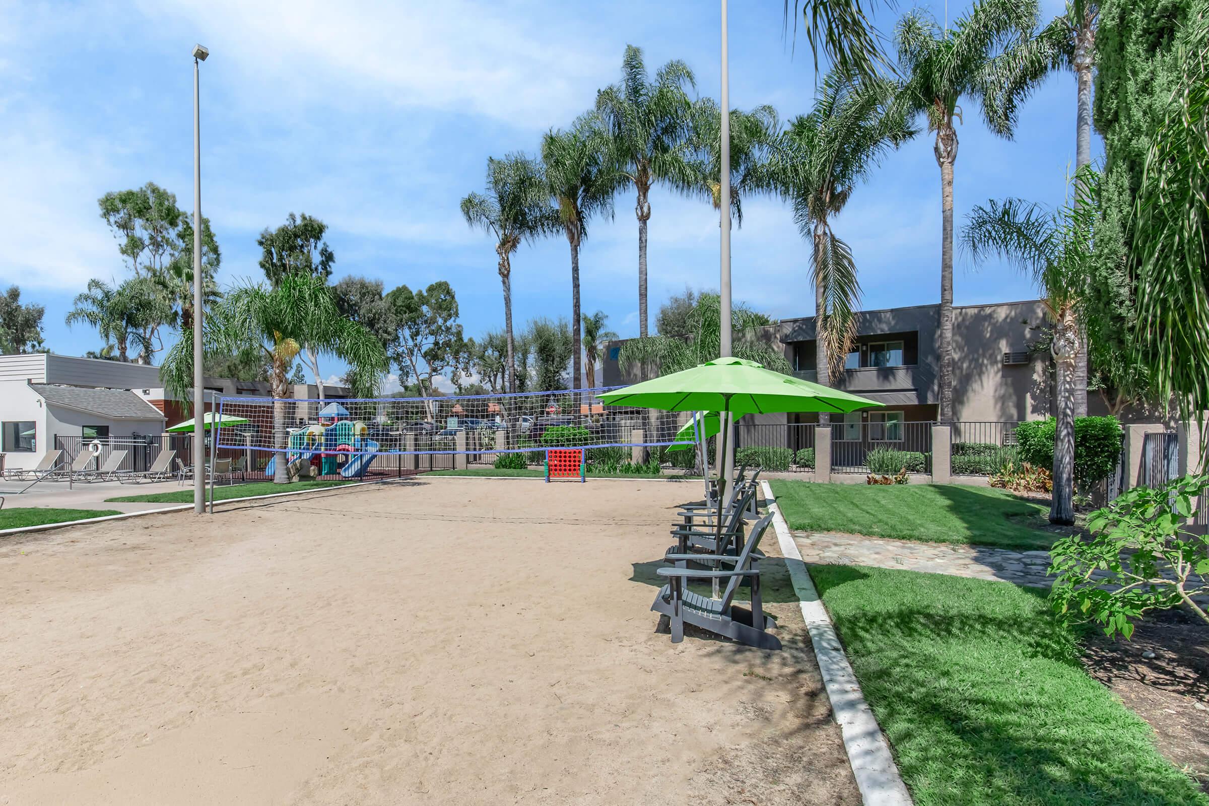A sandy volleyball court surrounded by palm trees and green umbrellas. Nearby, there are picnic tables and a playground area. The scene captures a sunny day in a recreational space, highlighting a relaxed outdoor environment.