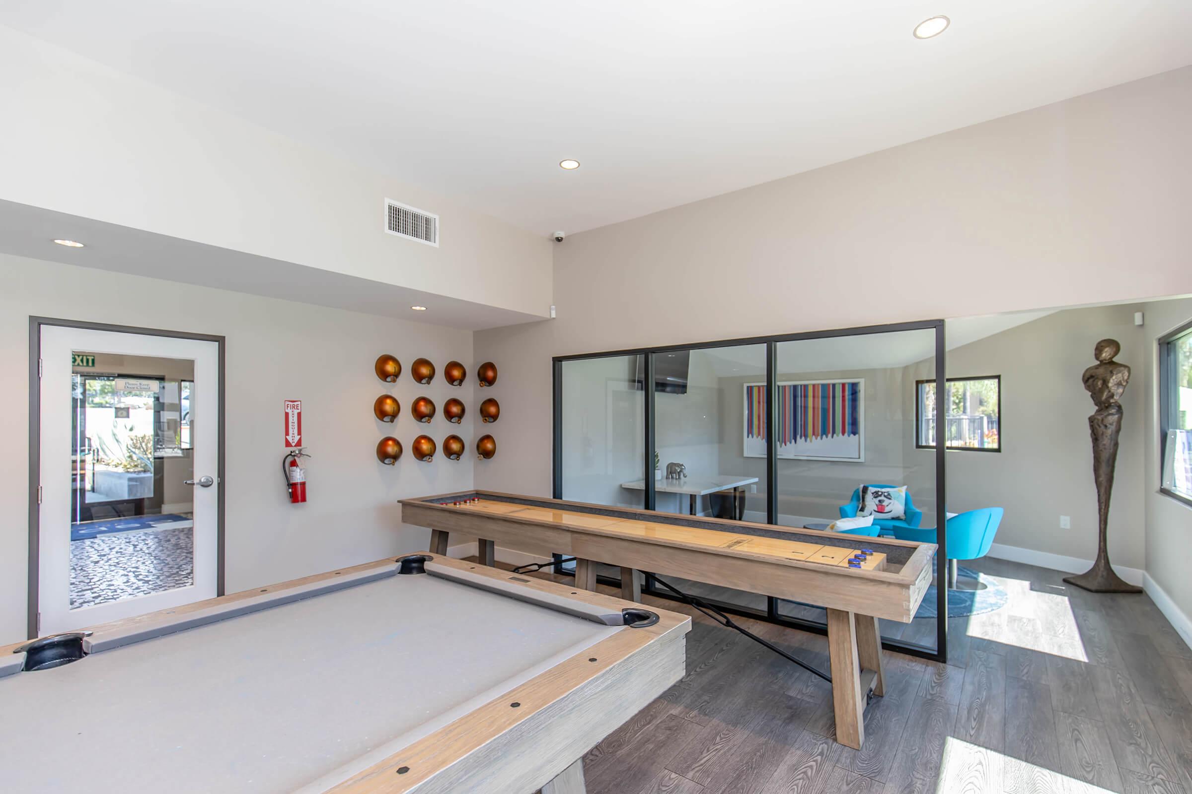 Interior of a modern recreational room showcasing a pool table and a shuffleboard table. A glass-enclosed office area is visible in the background, featuring colorful artwork and a blue chair. Sculptural decor adorns the wall, and ambient lighting enhances the inviting atmosphere.