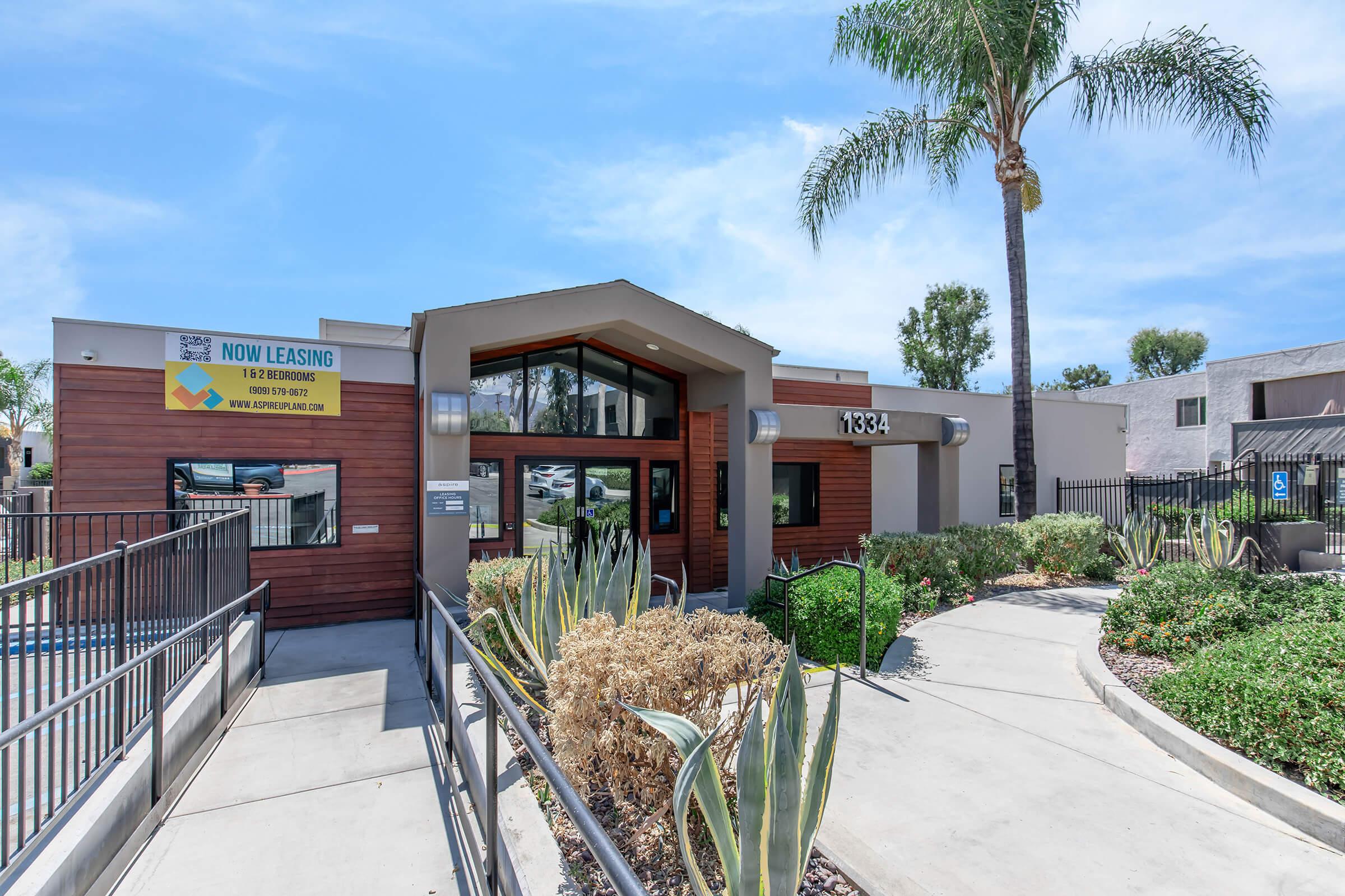 A modern leasing office building with a welcoming entrance, surrounded by landscaped greenery and palm trees. The sign above the door indicates that units are available for leasing. The building features large windows and a contemporary design, set against a clear blue sky.