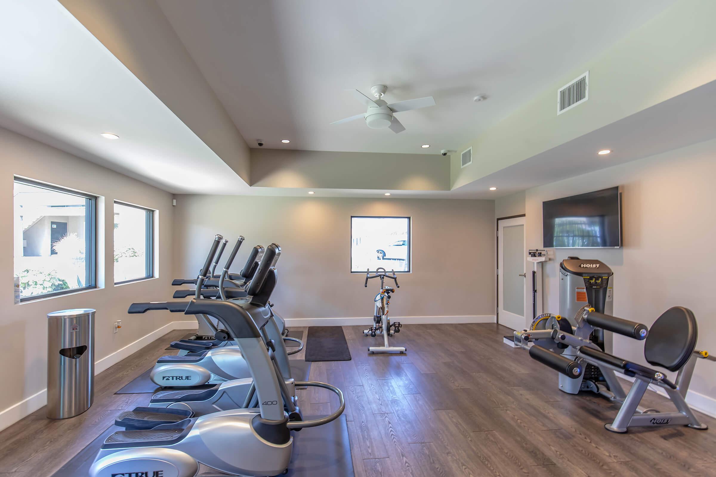 A modern fitness room featuring several exercise machines including treadmills and a stationary bike. There are large windows allowing natural light, a ceiling fan, a wall-mounted TV, and a trash bin. The flooring is wood-like, adding warmth to the space.