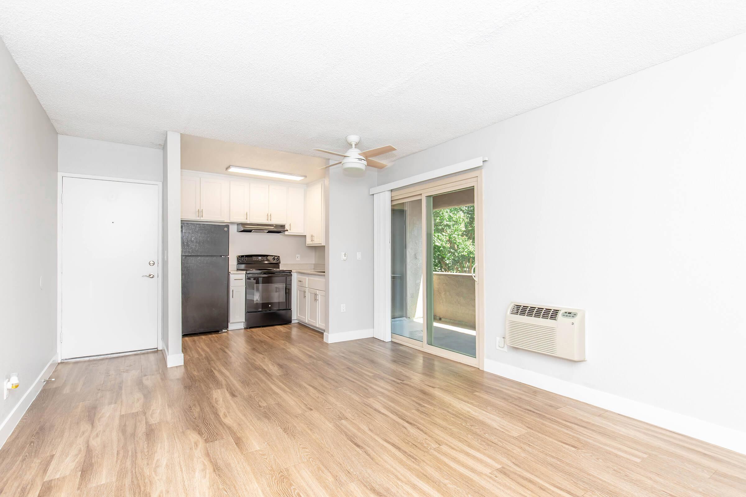 Spacious apartment interior featuring a light-colored wall, laminate flooring, a kitchen area with white cabinetry, stainless steel appliances, and a ceiling fan. A glass door leads to a small balcony or patio area. An air conditioning unit is mounted on the wall, providing a modern and inviting atmosphere.