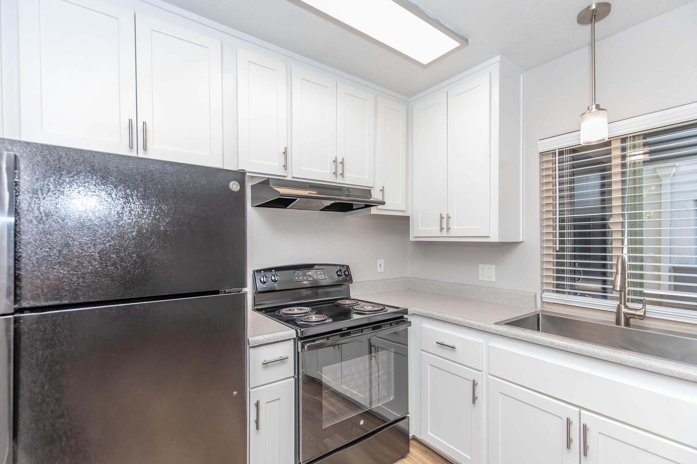 Modern kitchen featuring white cabinetry, a black refrigerator, and a stainless steel oven with a glass cooktop. There is a stainless steel sink under a window with blinds, and overhead lighting provides illumination. The countertops are light gray, offering a clean and bright look to the space.