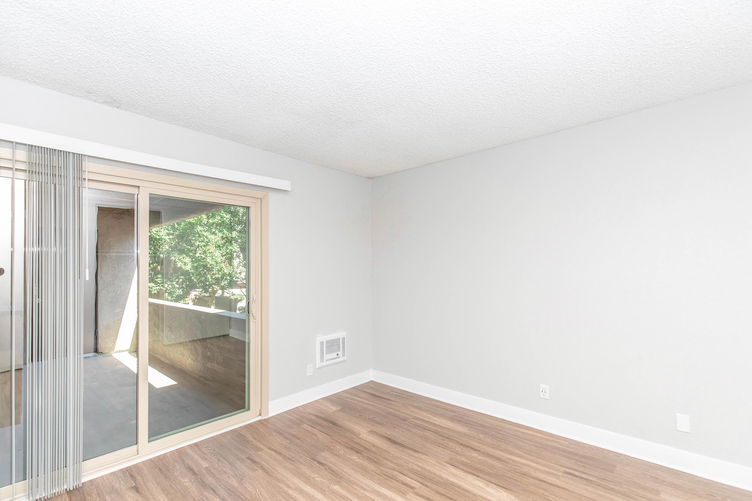 A light-filled room with a smooth, textured white wall and wooden flooring. Large sliding glass doors lead to a balcony, with vertical blinds partially closed. A small air conditioning unit is visible on the wall, and the overall space is uncluttered, providing a modern, minimalist aesthetic.