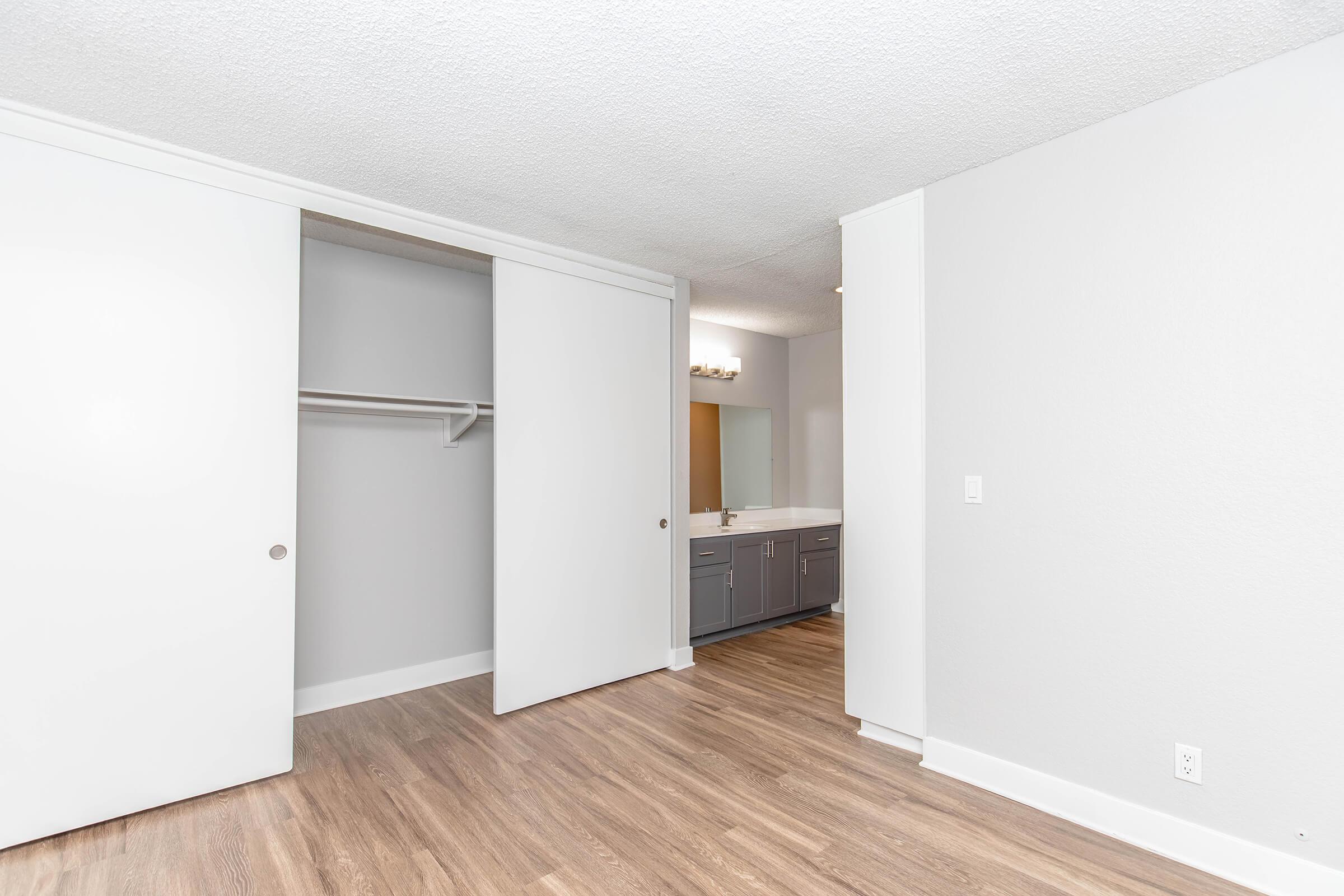 A modern, airy bedroom featuring light-colored walls and laminate flooring. It includes a spacious closet with sliding doors and a bathroom area visible in the background, showcasing a double sink and bright lighting. Natural light enhances the inviting atmosphere of the room.