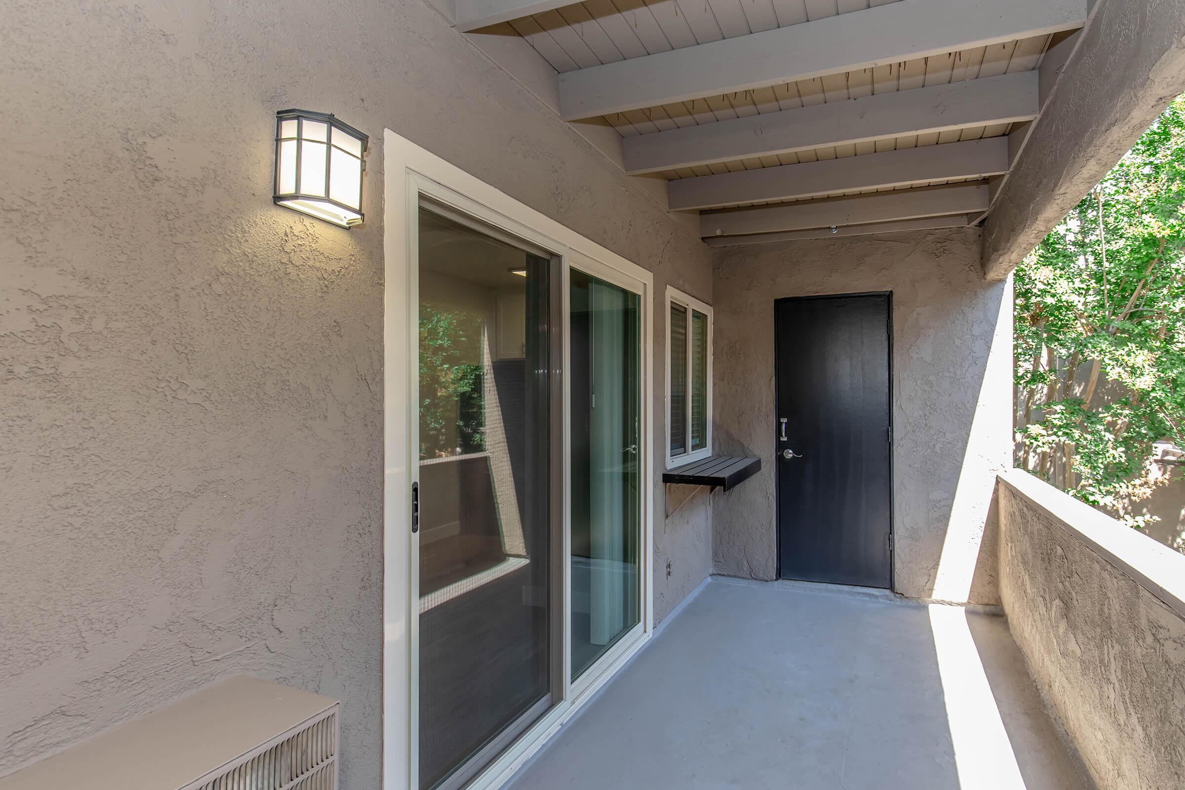 A balcony area with a light fixture, sliding glass doors, and a dark door. The walls are textured, and the ceiling features wooden beams. There is a small ledge along one wall, and the space is surrounded by greenery visible outside. The floor is concrete and well-lit.