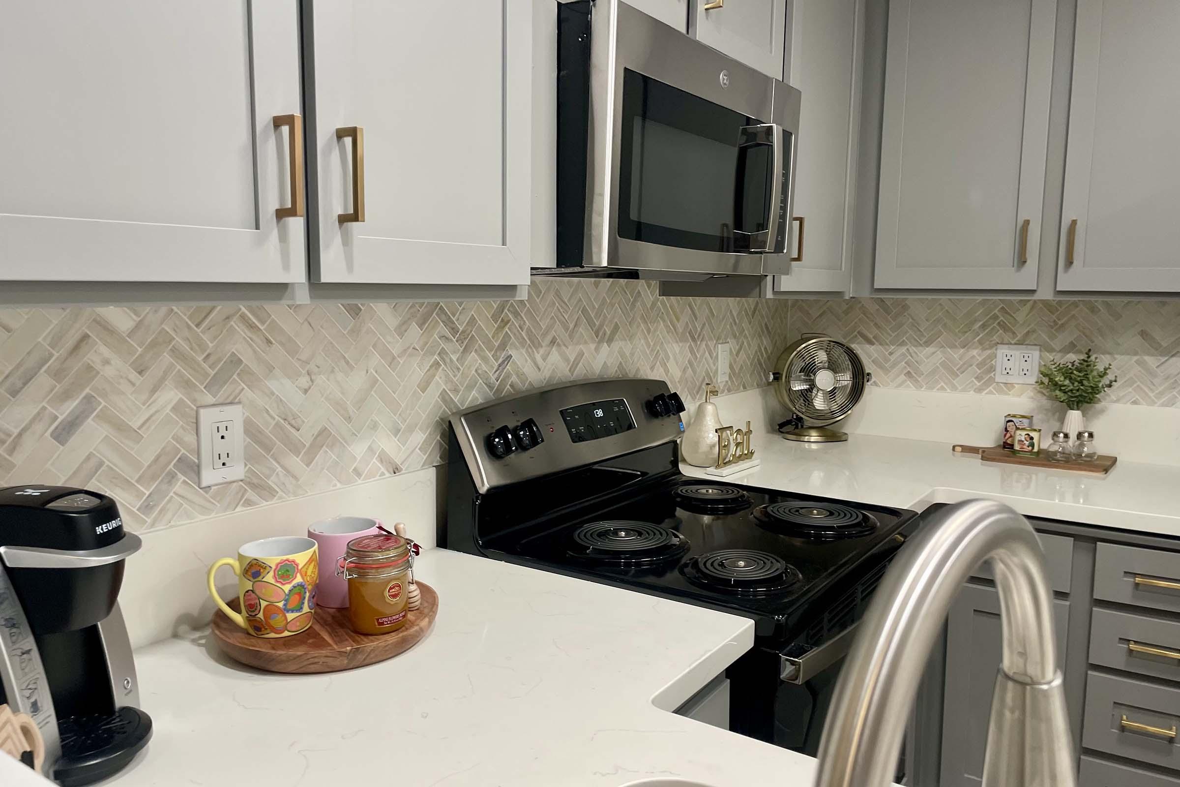 A modern kitchen featuring gray cabinets, a black stove, and an overhead microwave. The countertop includes a coffee maker, colorful mugs, and a round wooden tray. The backsplash has a herringbone pattern in neutral tones, and a decorative fan sits on the counter. The overall design is bright and inviting.