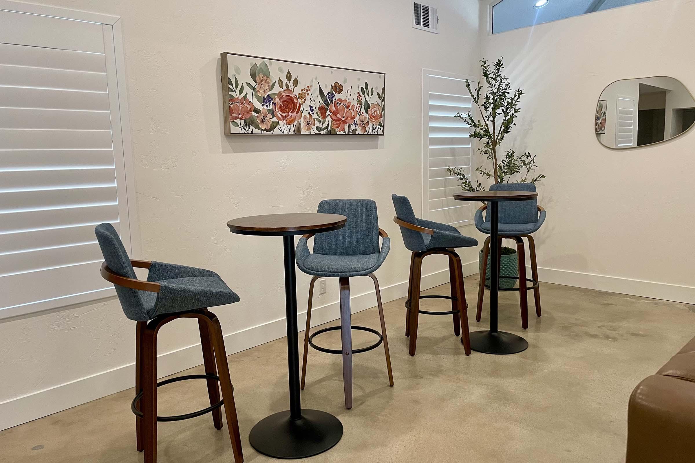 A modern interior space featuring a small round table with four blue upholstered bar stools. The wall includes a floral artwork, and there are decorative plants nearby. White shutters provide natural light, and the floor is a light polished concrete.