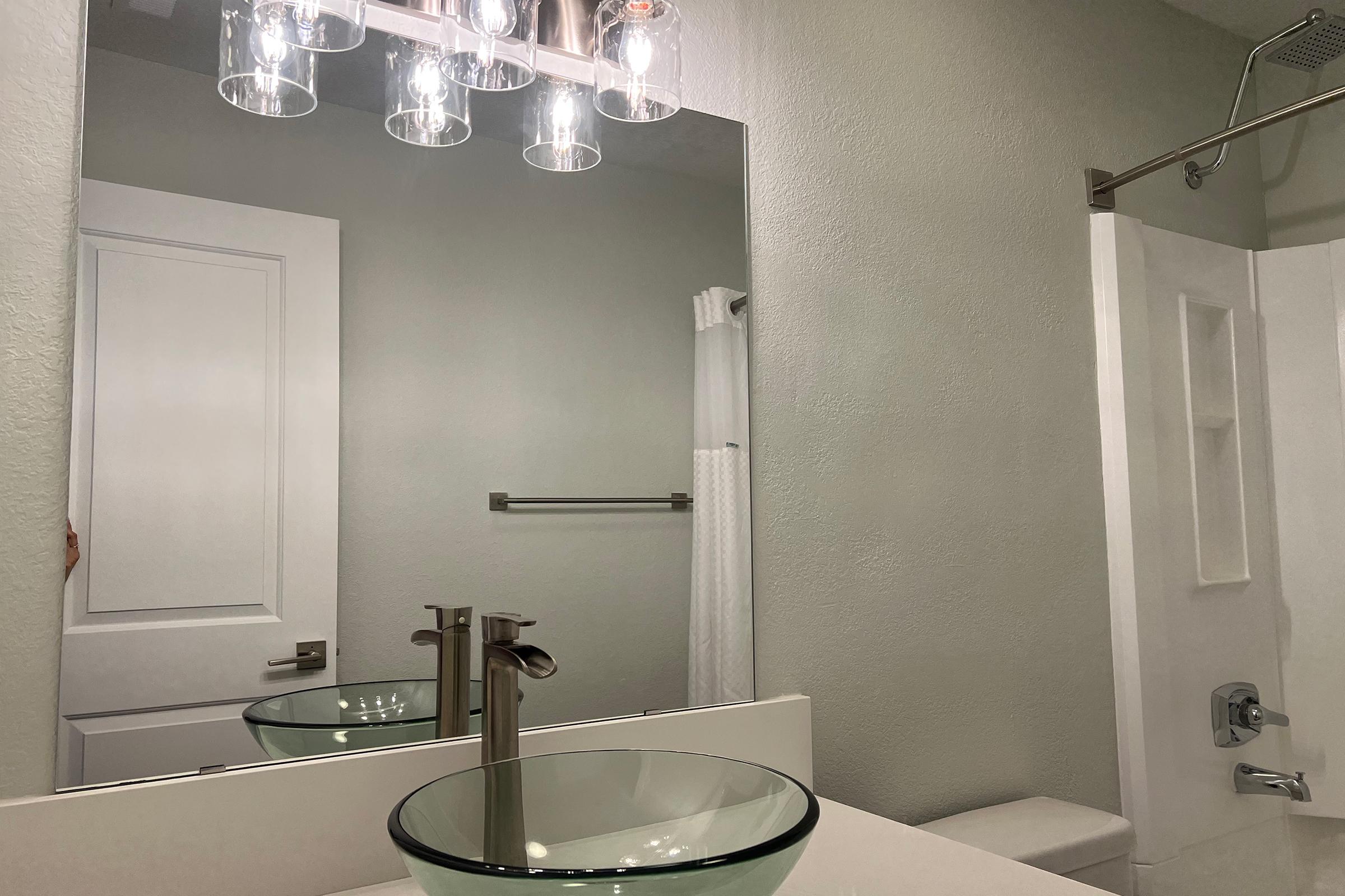 A modern bathroom featuring a stylish glass sink, a large wall-mounted mirror with five light fixtures above, a shower with a glass door, and neutral-colored walls. A towel rack is visible, along with a white shower curtain.