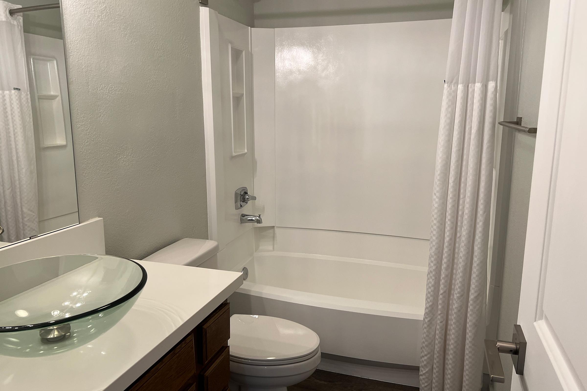 A clean, modern bathroom featuring a white tub with a shower, a glass vessel sink on a light countertop, wooden cabinetry, and a mirror. The walls are painted a soft gray, and there is a white shower curtain. The floor has a neutral tone, adding to the contemporary feel.