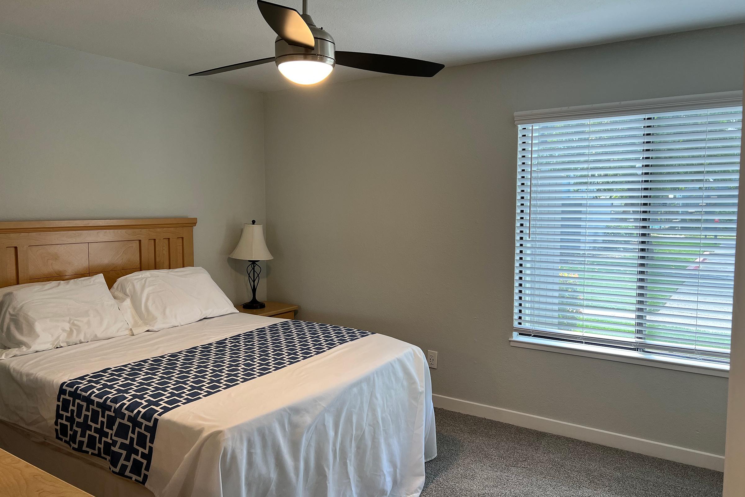 A neatly arranged bedroom featuring a bed with white linens and a blue accent blanket. There is a bedside lamp on a wooden nightstand beside the bed. A ceiling fan is overhead, and a window with blinds offers natural light, illuminating the light gray walls and carpeted floor.