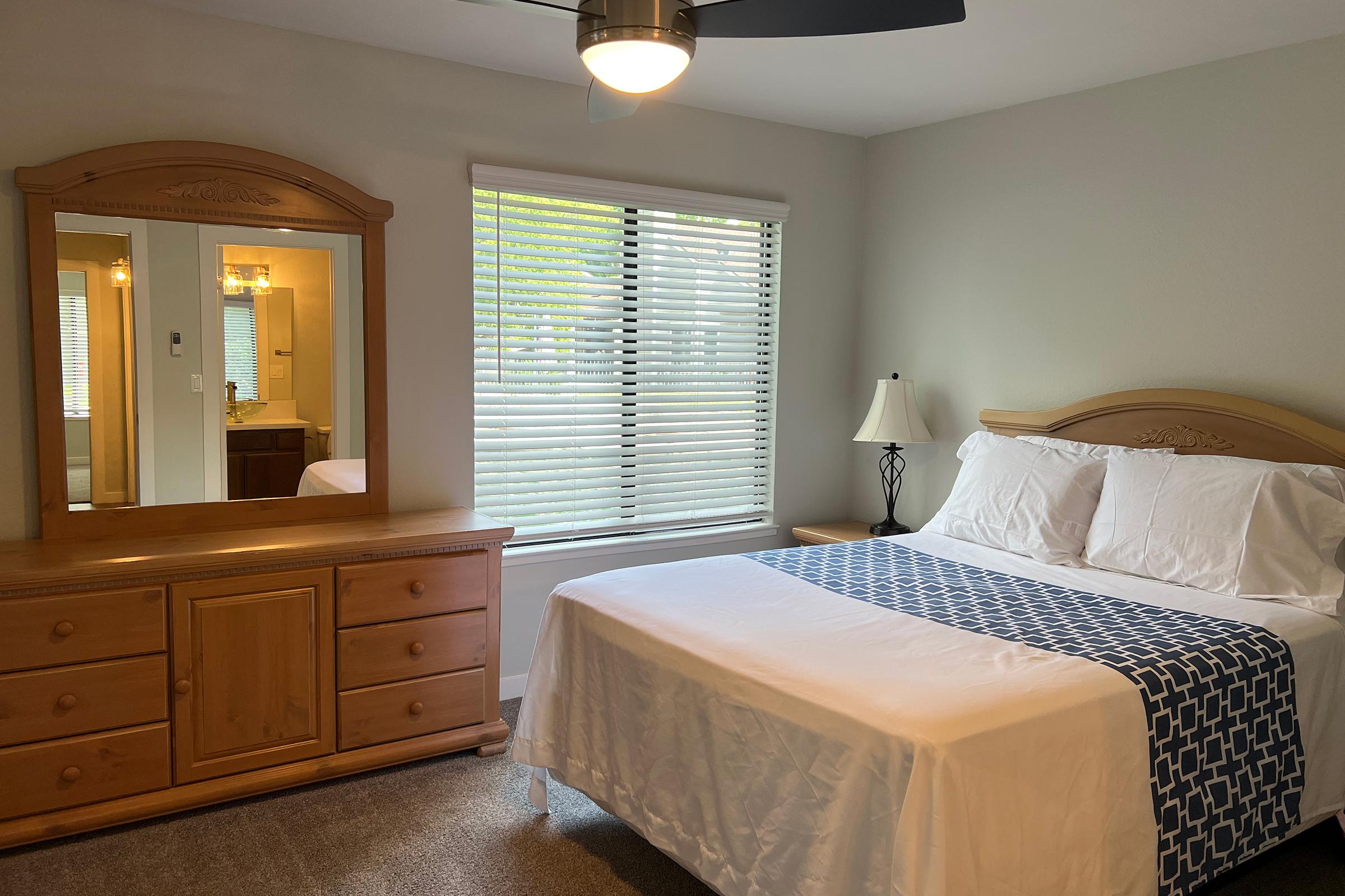 Brightly lit bedroom featuring a queen-sized bed with white linens and a blue patterned throw. A wooden dresser with a matching mirror sits beside the bed. A lamp stands on a nightstand, and a bathroom can be seen through an open door with light-colored walls and blinds on the window.