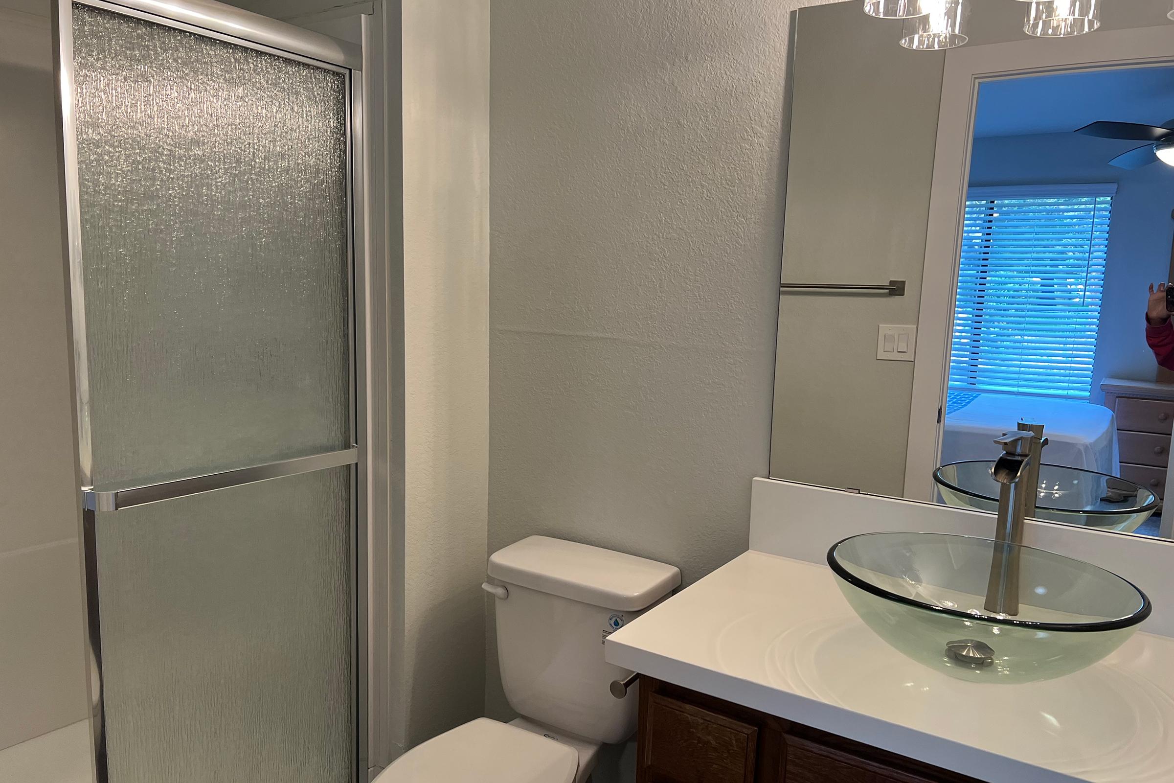 A modern bathroom featuring a glass shower enclosure, a white toilet, and a stylish sink with a round glass basin. A large mirror is mounted above the sink, and the room is softly lit, with natural light coming through a window that is partially covered by blinds.