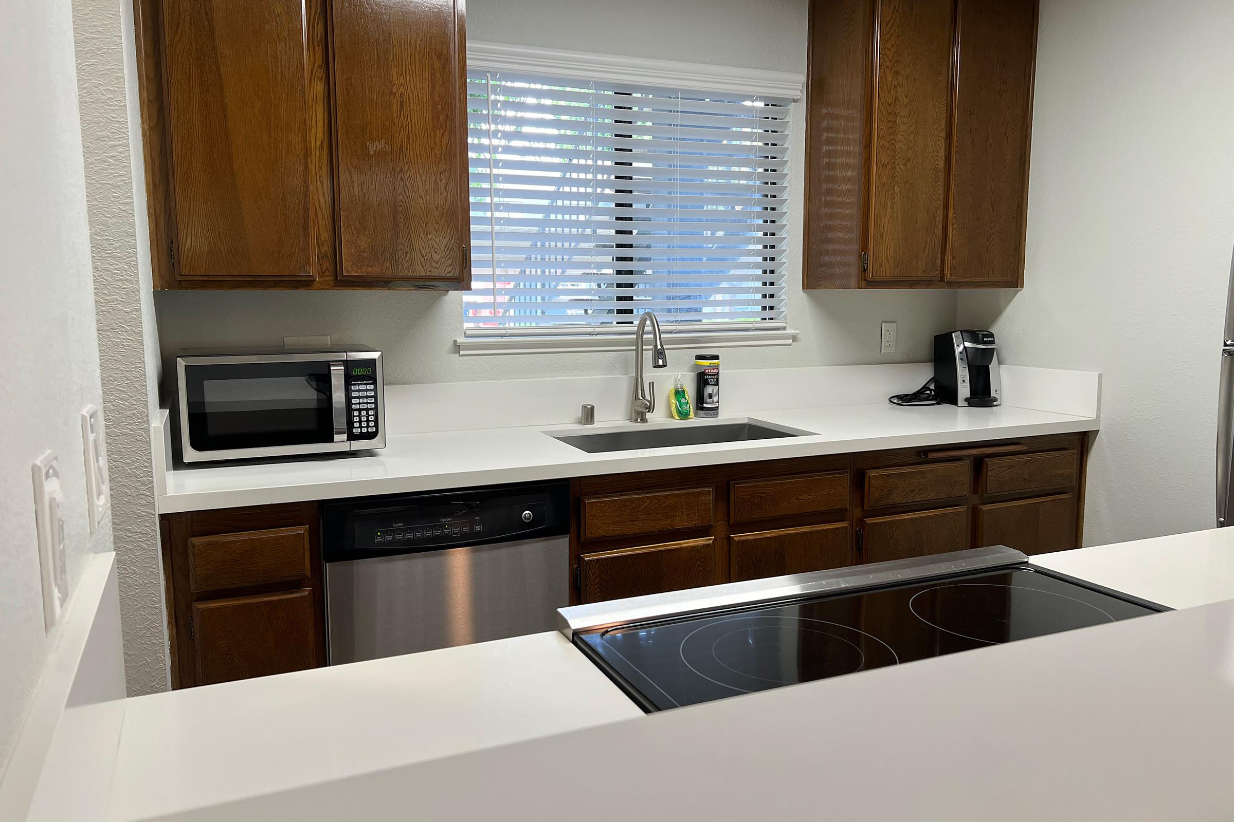 Modern kitchen with wooden cabinets, a microwave, a stainless steel dishwasher, and a sink. The countertop is white and smooth, with an induction cooktop visible. There are some kitchen appliances and utensils, and a window with blinds allows natural light to enter.