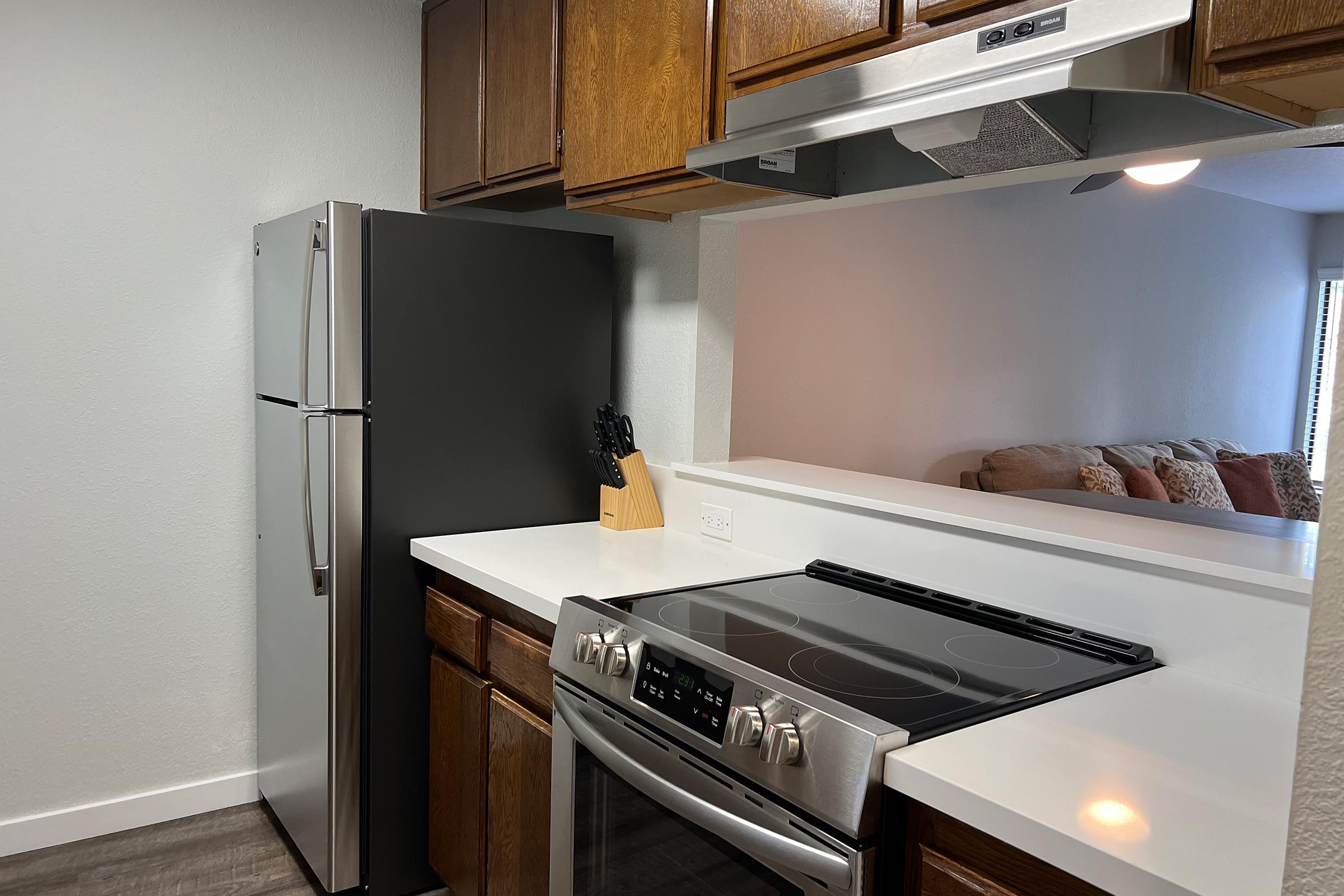 A modern kitchen featuring a stainless steel refrigerator, a sleek black stove with a five-burner cooktop, and wooden cabinets. The countertop is white and smooth, with a knife block neatly placed beside the stove. In the background, a cozy living area with a couch is visible.