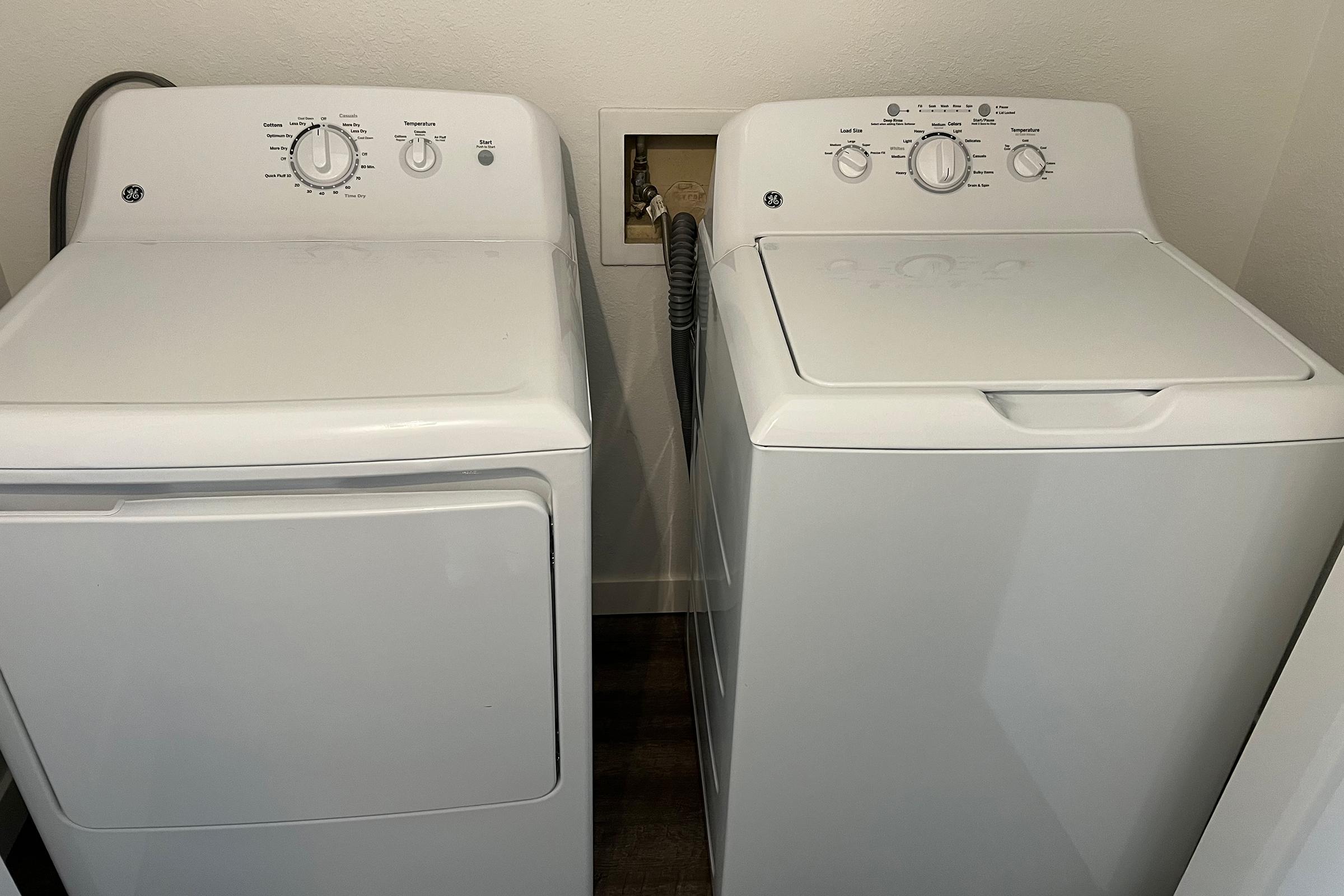 Two white laundry appliances: a front-loading dryer on the left and a top-loading washing machine on the right. The dryer features control knobs and has a vent connected to the wall, while the washing machine has a lid on top and knobs for selecting cycles. The appliances are located in a small laundry area.