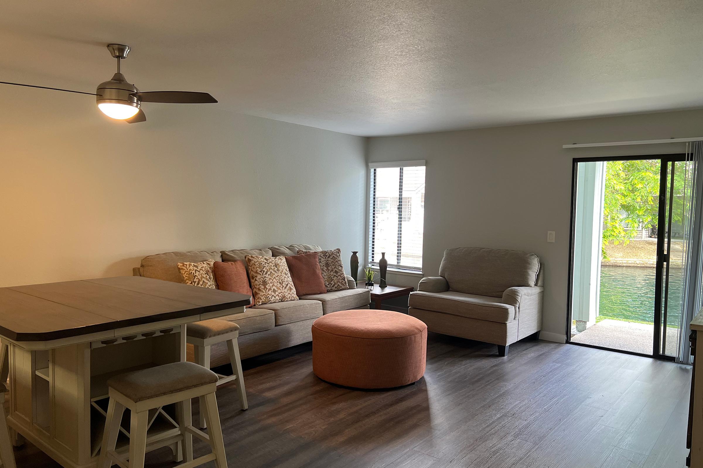 A bright living room featuring a beige sectional sofa with decorative pillows, a round orange ottoman, and a comfortable chair. A dining table with stools is visible, along with a sliding glass door leading to an outdoor area with greenery. A ceiling fan provides a cozy atmosphere.