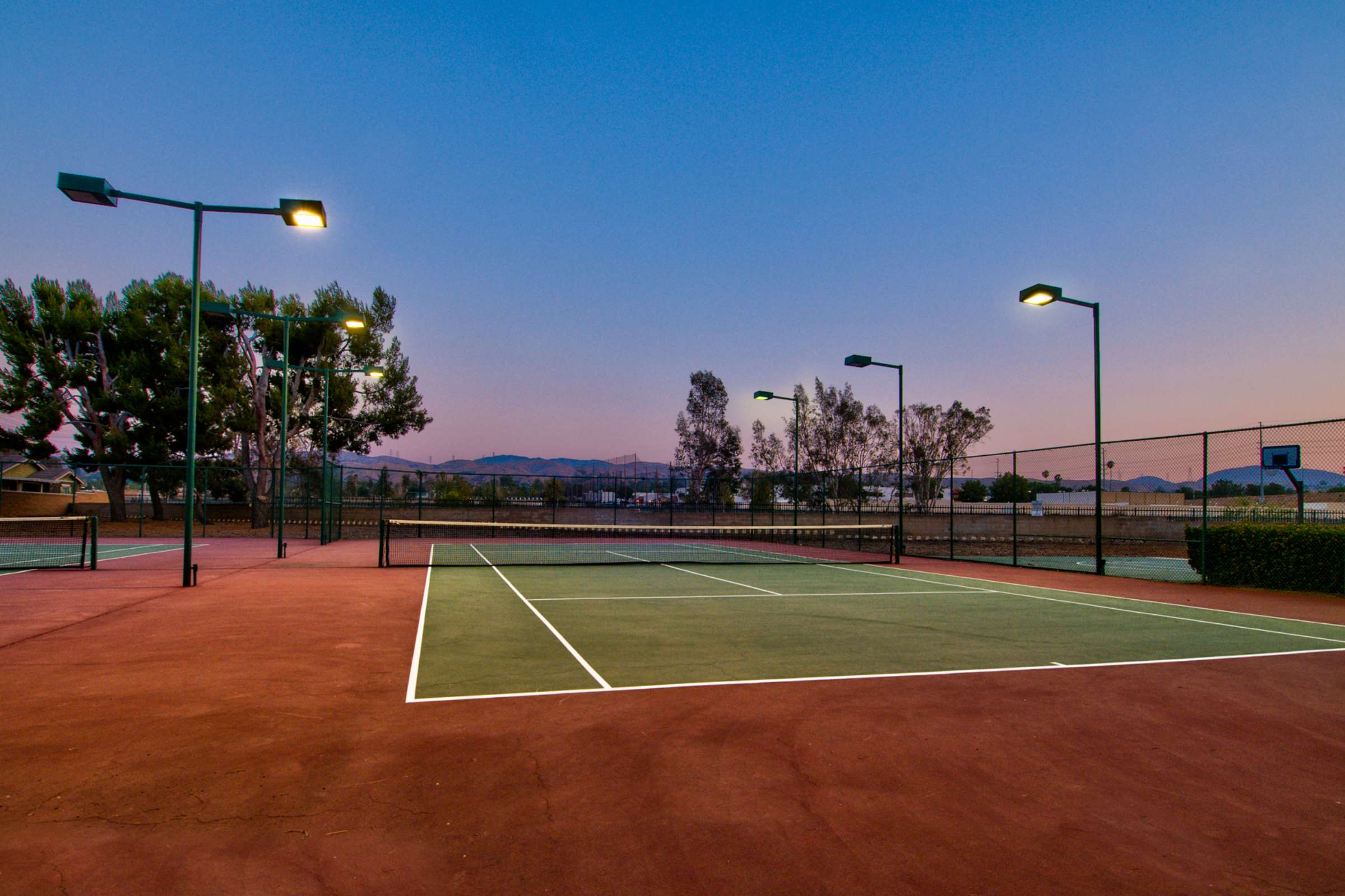 An empty tennis court at dusk, with floodlights illuminating the area, surrounded by trees and mountains in the background.