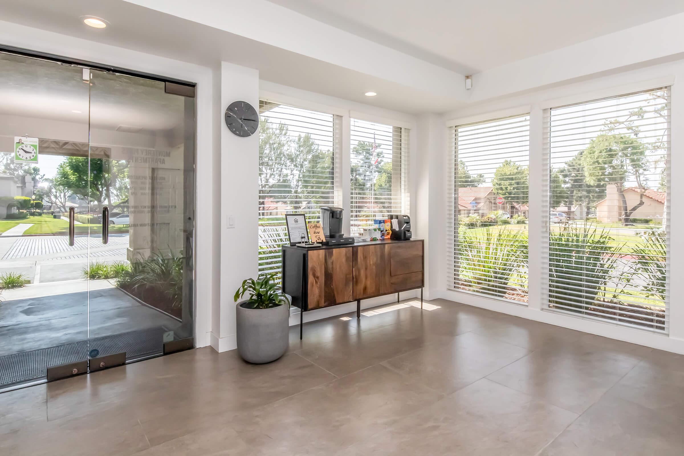 A modern interior space featuring a large glass entrance door, contemporary furnishings, and natural light streaming through large windows with white blinds. A decorative wooden cabinet holds various items, and plants add a touch of greenery. The view outside shows a landscaped area.