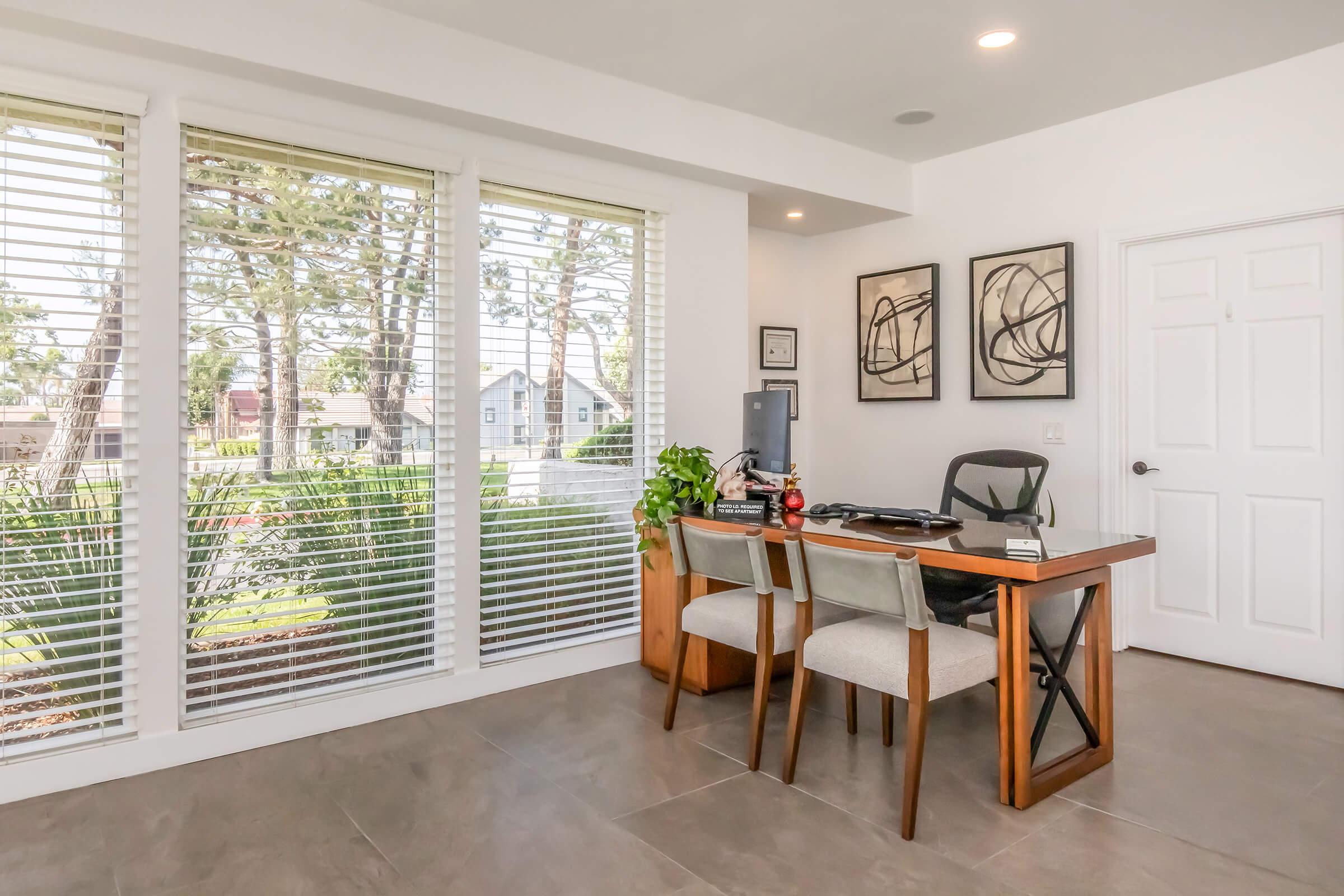 A modern home office featuring a wooden desk with two chairs, a small plant on the desk, and art prints on the walls. Large windows with blinds let in natural light, offering a view of greenery outside. The room has a clean, minimalist design with tile flooring.