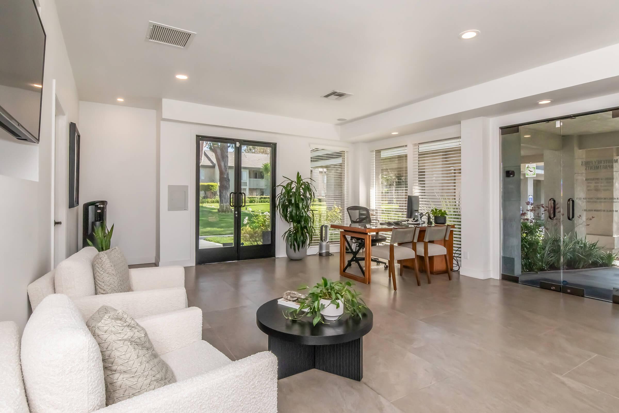 Modern office lounge area featuring two light-colored sofas, a round black coffee table with plants, and a glass door leading to an outdoor space. A desk and chair are visible in the background, alongside large windows allowing natural light to flood the room. Lush greenery can be seen outside.