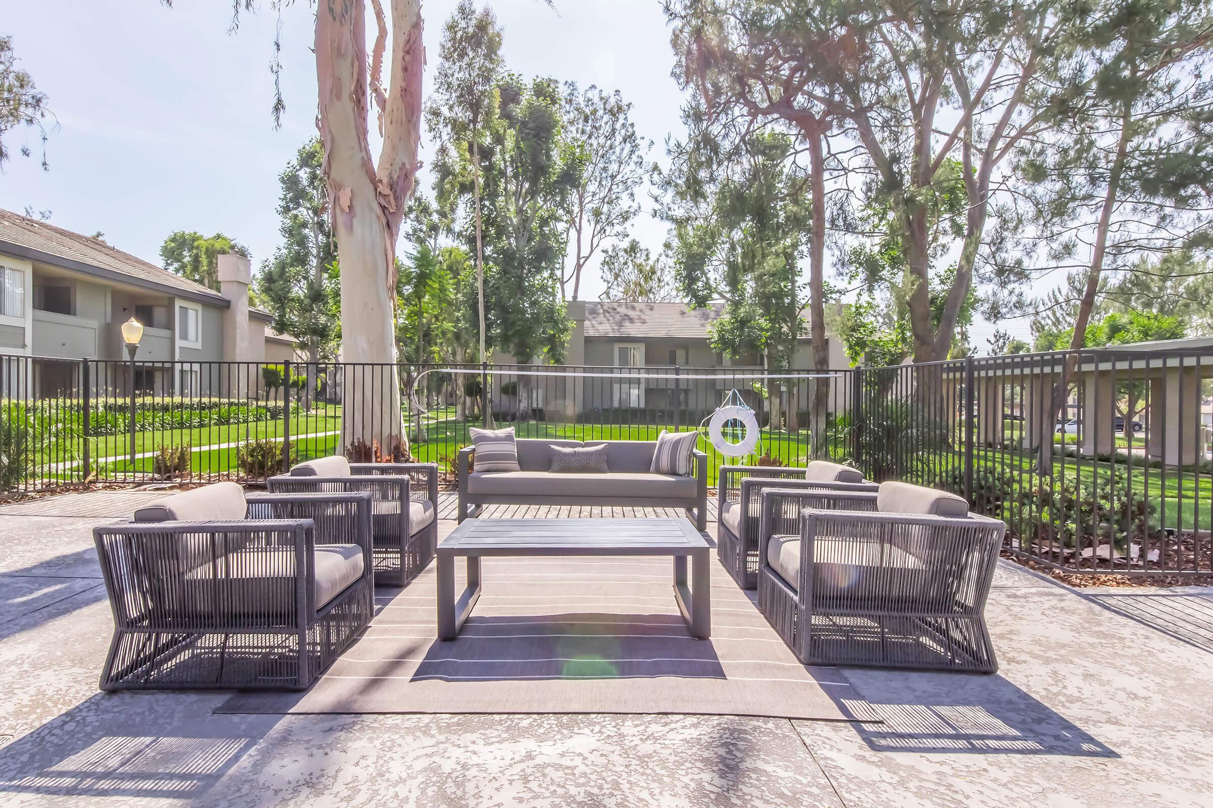 A cozy outdoor seating area featuring a modern gray table surrounded by four matching armchairs. The space is situated in a well-maintained garden with trees and greenery in the background. A fence encloses the area, creating a private atmosphere for relaxation and gatherings.