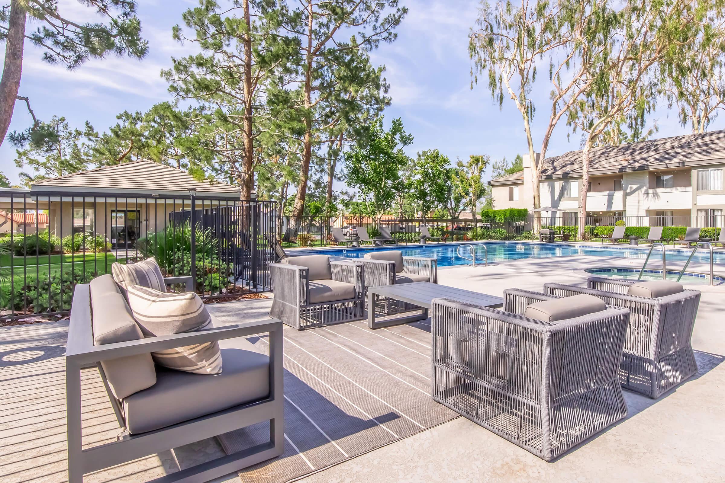 Outdoor pool area featuring a sunny atmosphere with a large swimming pool surrounded by trees. A stylish seating arrangement of modern chairs and a table is placed on a deck beside the pool. Reflections of greenery are visible in the water, creating a relaxing, inviting environment.