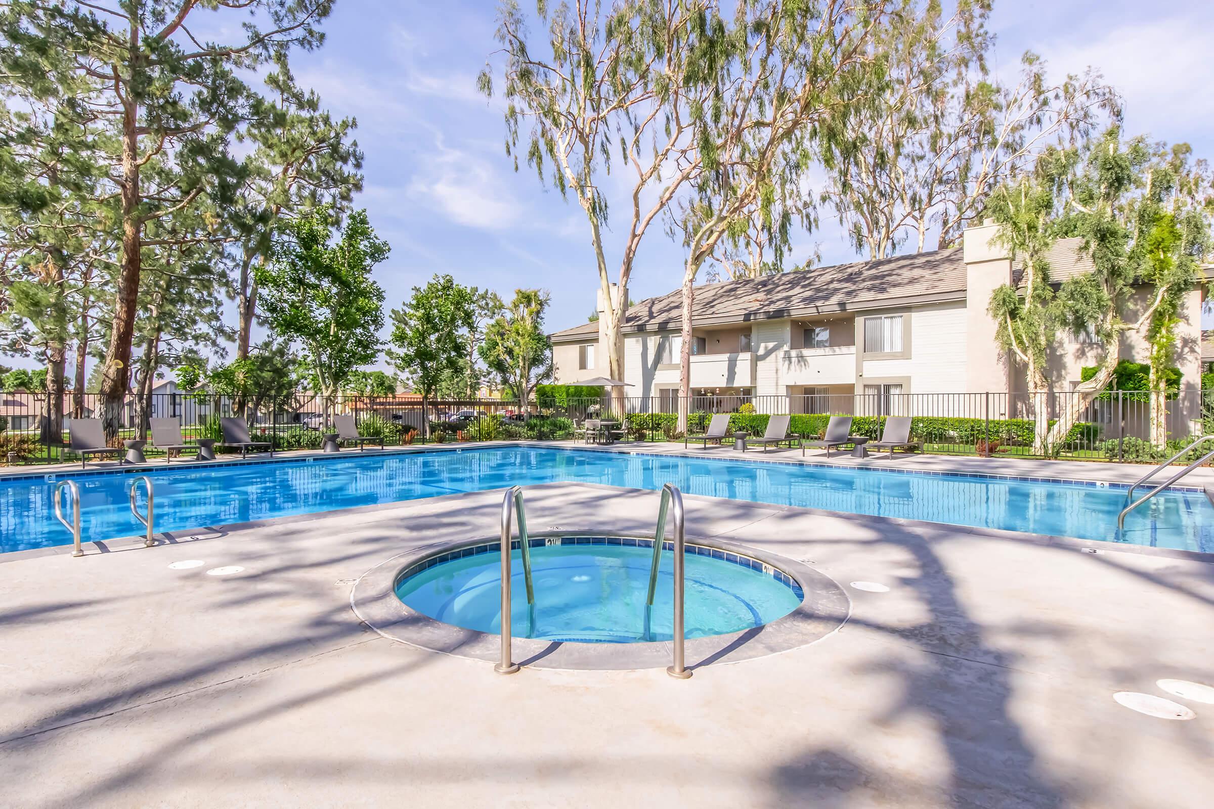 A serene outdoor swimming pool area featuring a larger pool and a smaller spa. Surrounding the pool are tall trees and well-maintained landscaping, with lounge chairs lining the edges. In the background, there's a multi-story building with several balconies, creating a relaxing atmosphere.