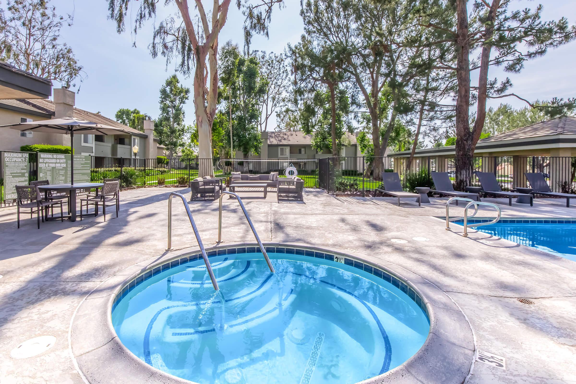 A beautifully landscaped outdoor area featuring a swimming pool and a hot tub, surrounded by lounge chairs and seating arrangements. Lush trees and grass provide a serene ambiance, with modern apartment buildings visible in the background. Clear blue sky enhances the relaxing atmosphere.
